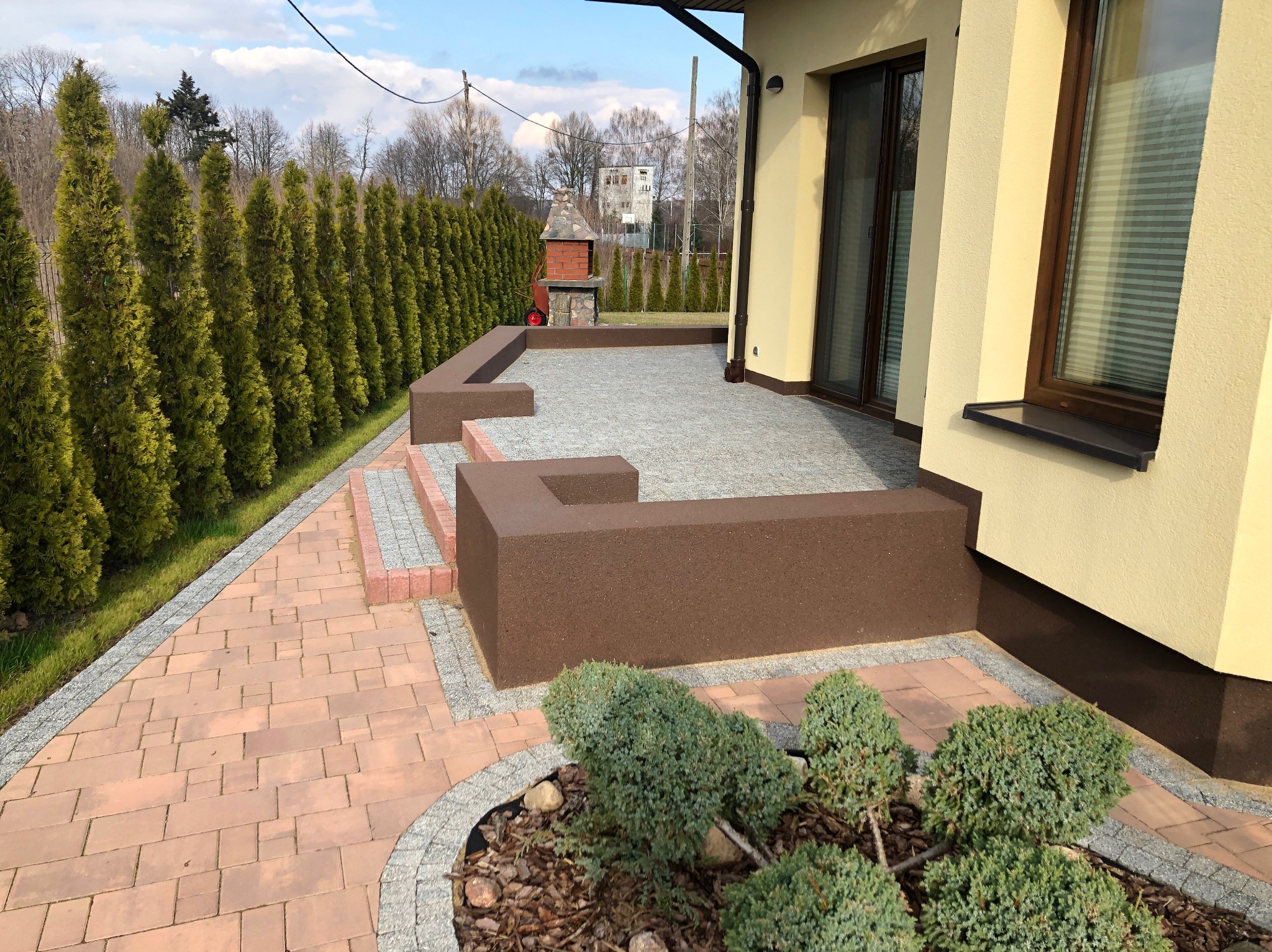 Brukowa ścieżka z obrzeżami prowadzi do tarasu z grillem, otoczona tujami i ozdobnymi krzewami. Elementy małej architektury w harmonijnej kolorystyce.