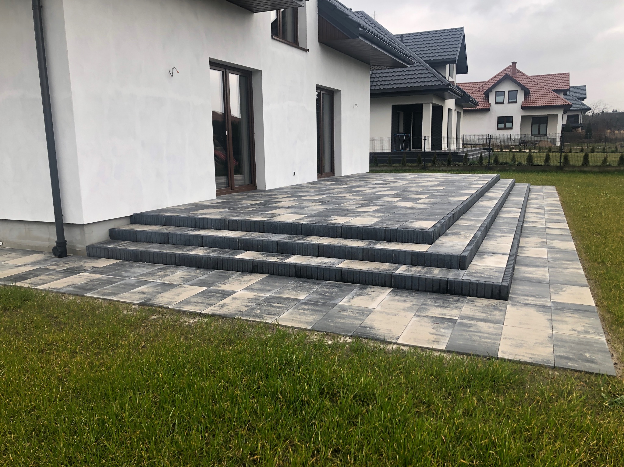 Nowoczesny taras z kostki brukowej w odcieniach szarości, prowadzący do białego domu z ciemnymi oknami. Widok na zadbany trawnik i sąsiednie domy.