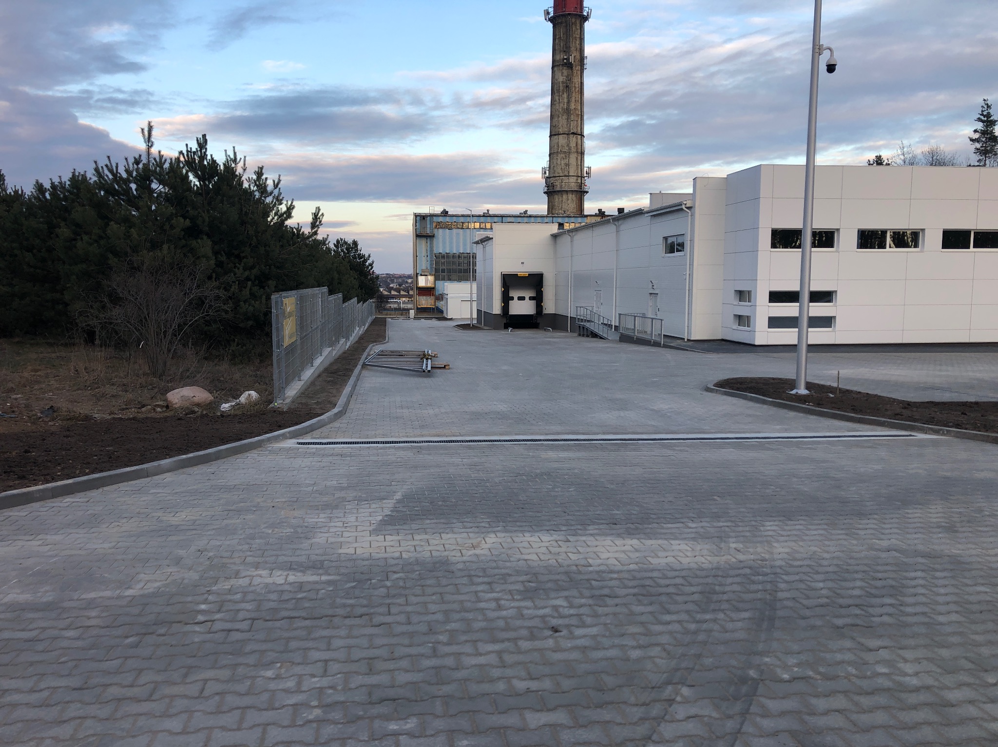 Szara kostka brukowa ułożona na podjeździe w Starych Kupiskach, z widocznymi krawężnikami i kratkami odpływowymi. W tle budynek przemysłowy i komin.