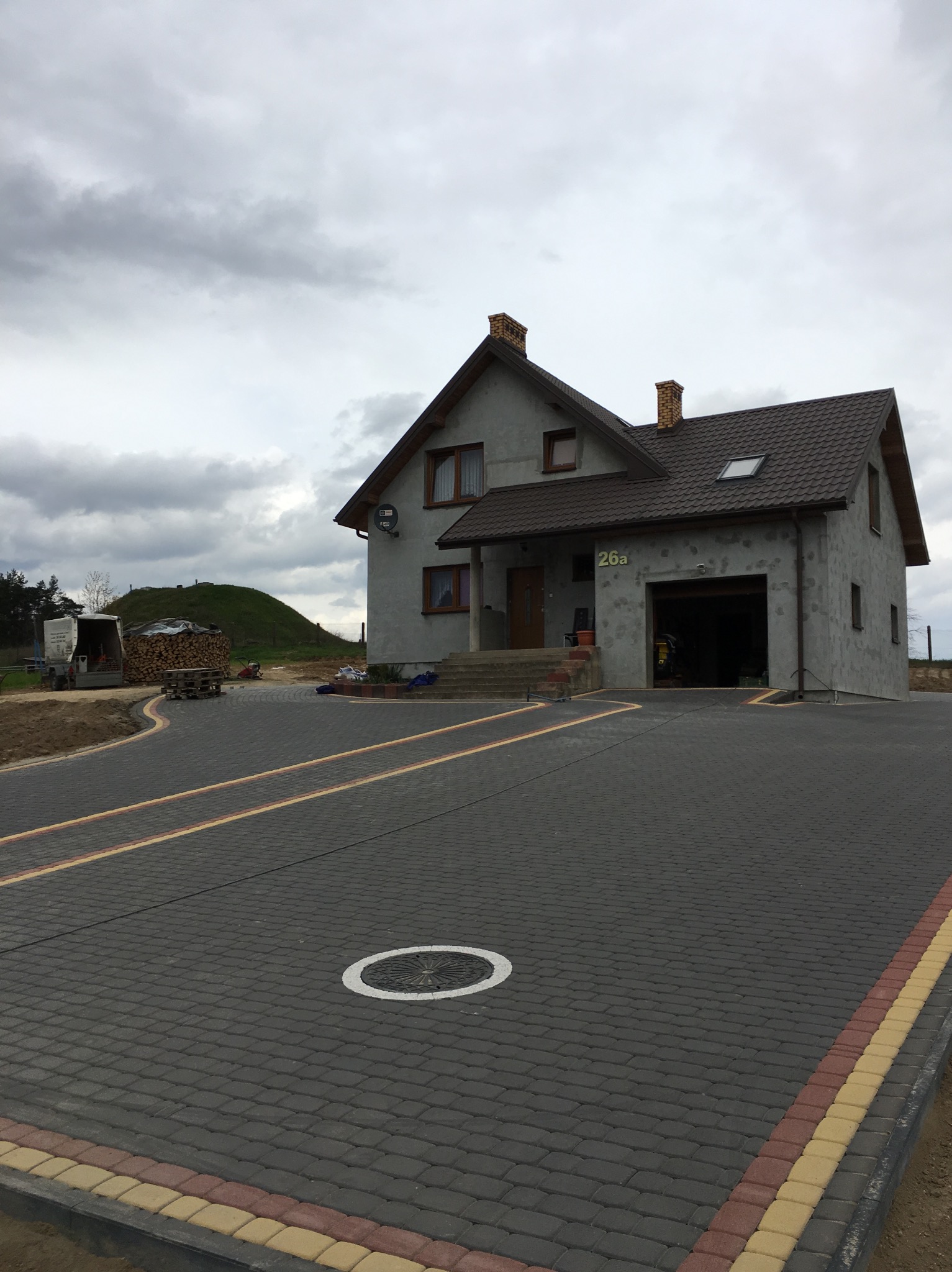 Ułożona, grafitowa kostka brukowa z obrzeżami w odcieniach beżu i czerwieni prowadzi do domu z garażem. Widoczny właz kanalizacyjny. W tle hałda ziemi i zapas drewna.