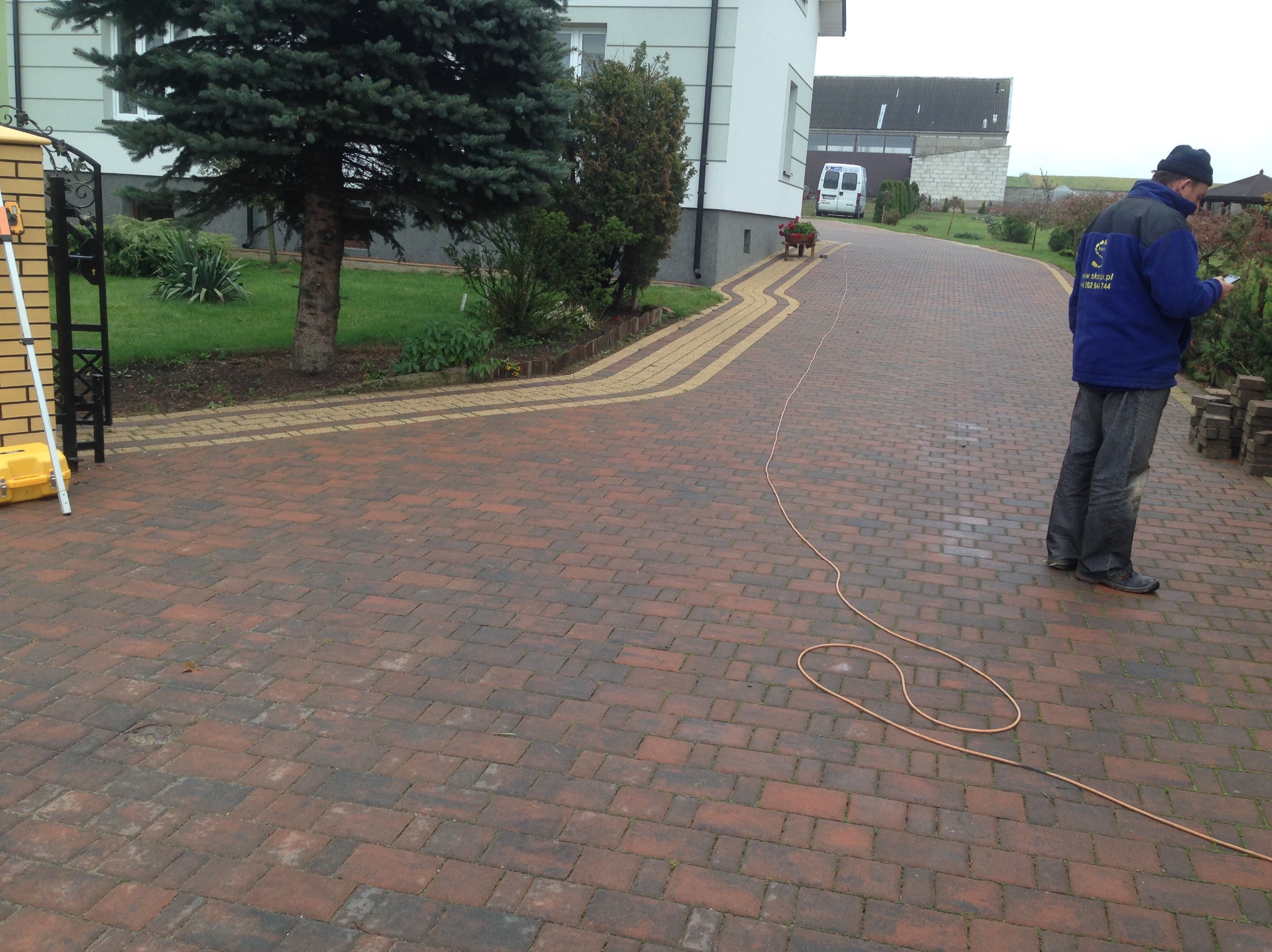 Kaskadowa ścieżka z kostki brukowej w odcieniach czerwieni i beżu, wijąca się obok trawnika z drzewami. W tle budynek mieszkalny i gospodarczy, a także pracownik z telefonem.