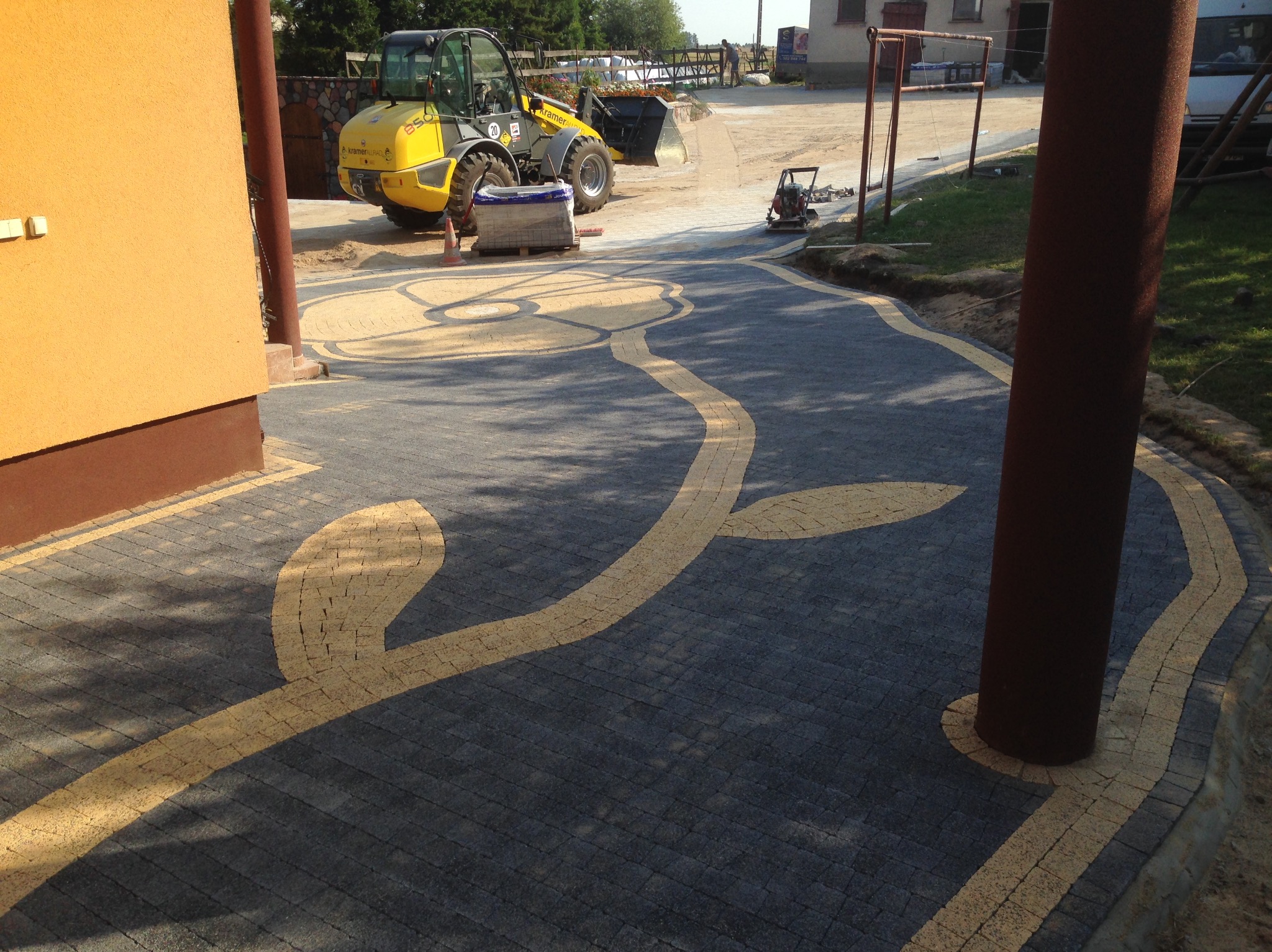 Ukończona ścieżka z kostki brukowej w odcieniach szarości i beżu, ułożona we wzór kwiatowy. W tle maszyna budowlana i ubijarka.