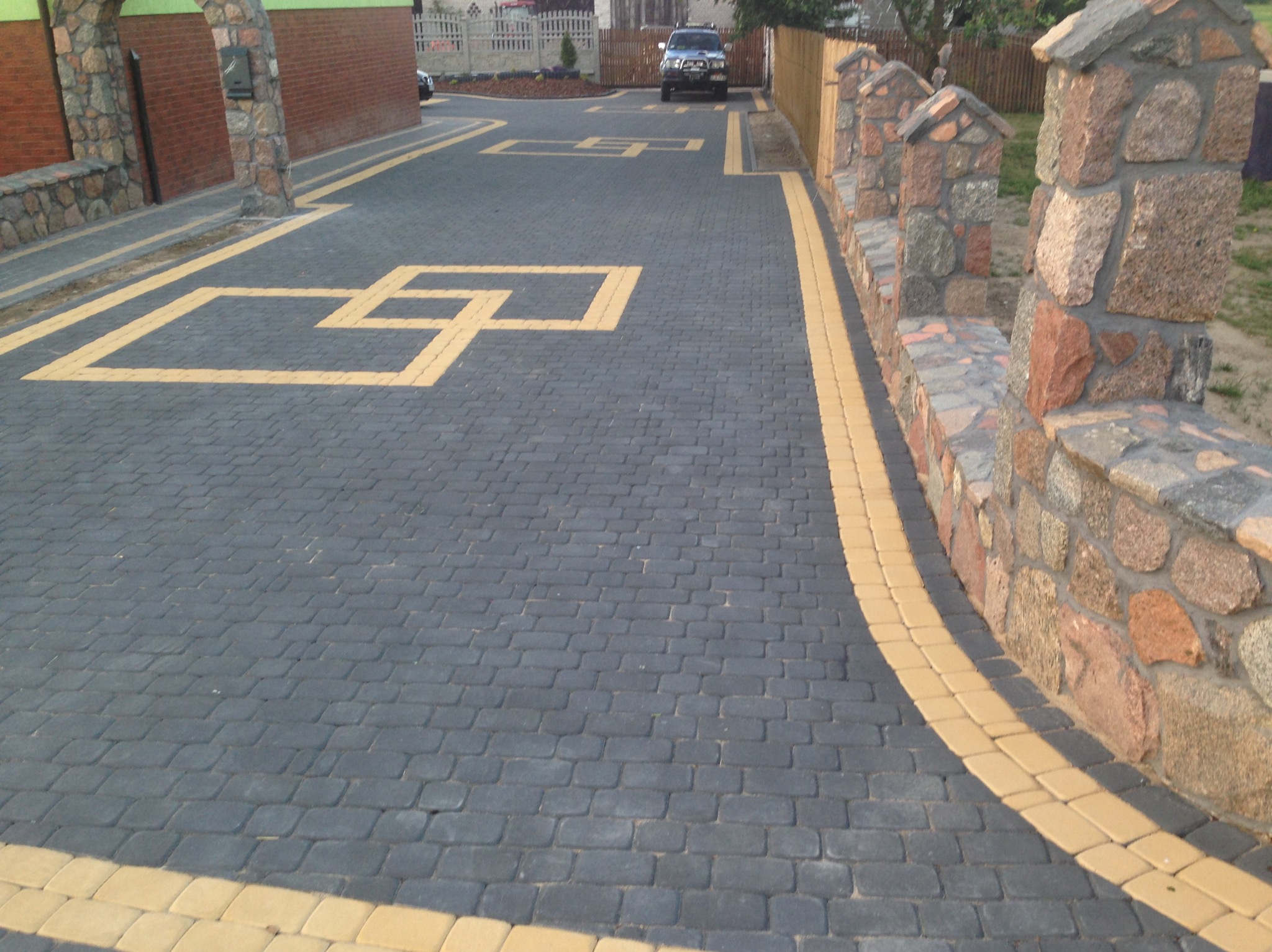 Ułożona, grafitowa kostka brukowa z żółtymi akcentami tworzącymi geometryczny wzór. Kamienny murek oddziela posesję. W tle zaparkowany samochód.