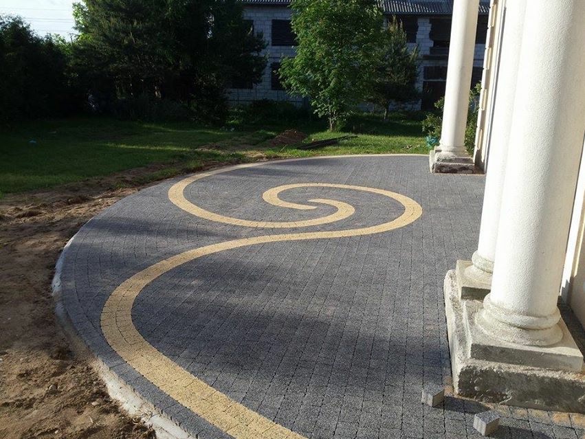 Szara kostka brukowa z żółtym wzorem spirali, otoczona kolumnami. Widok na podjazd z kostki, zieleń w tle. Estetyczne wykończenie terenu.