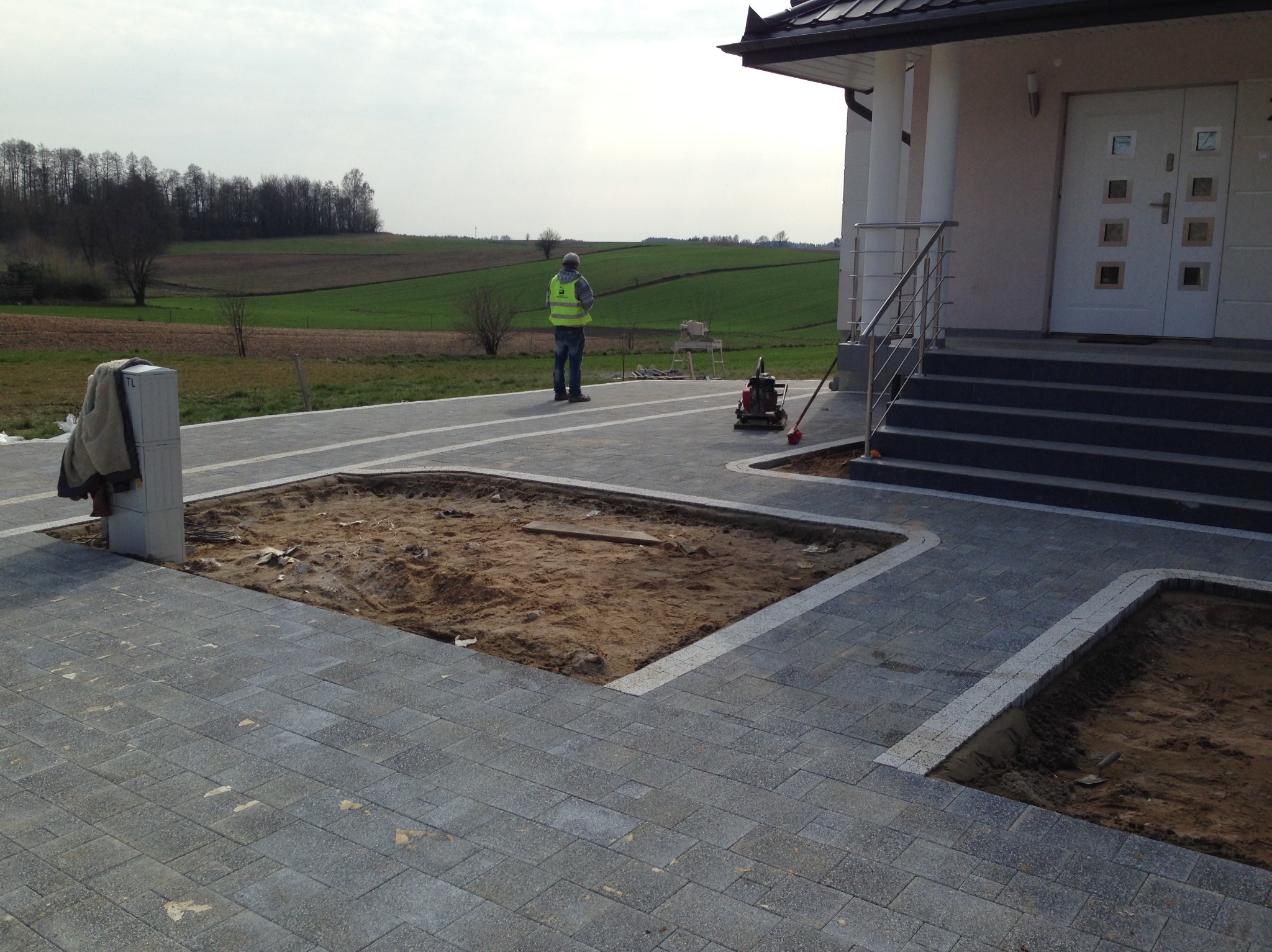 Fragment posesji z częściowo ułożoną kostką brukową, przygotowane miejsca pod nasadzenia, pracownik w kamizelce odblaskowej i zagęszczarka w tle.