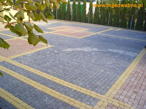 Szara kostka brukowa z żółtymi i brązowymi akcentami tworząca geometryczny wzór na podjeździe, z zielonymi tujami w tle i fragmentem drzewa w lewym górnym rogu.