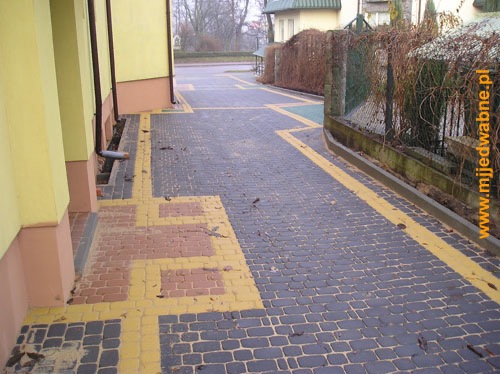 Ułożona kostka brukowa wzdłuż budynku z żółtymi akcentami, tworząca wzór. Widoczny fragment ogrodzenia i rynna. Kostka w odcieniach szarości i czerwieni.
