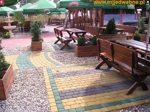 Kolorowa kostka brukowa tworzy ścieżkę w ogrodzie restauracji. Drewniane ławki i stoliki, donice z roślinami, żwirowe alejki. Widok na zewnątrz lokalu.