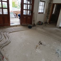 Serhii Rakomin - Wnętrze w trakcie remontu z ułożonymi rurami instalacji podłogowej na surowym betonie. Widoczne otwarte drzwi i okno. Wiadro i narzędzia w tle.