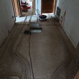 Serhii Rakomin - Korytarz w trakcie prac elektrycznych: liczne rury instalacyjne ułożone wzdłuż ścian, sprzęt elektryczny na podłodze, widoczne kable i narzędzia. Surowy stan wykończenia.