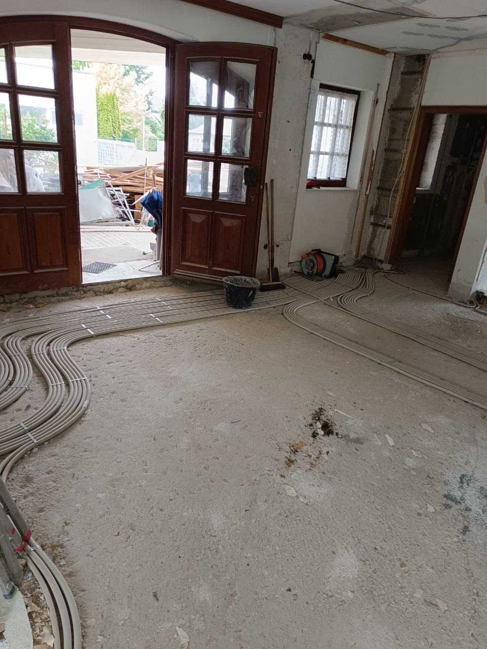 Wnętrze w trakcie remontu z ułożonymi rurami instalacji podłogowej na surowym betonie. Widoczne otwarte drzwi i okno. Wiadro i narzędzia w tle.
