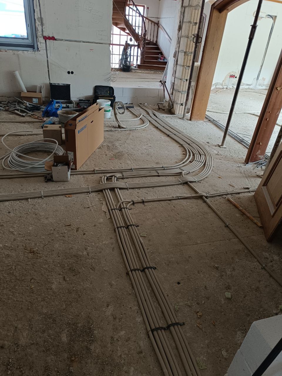 Rozprowadzone kable elektryczne w szarych rurkach na surowej podłodze w trakcie remontu domu. Widoczne skrzynki, narzędzia i schody w tle. Instalacja elektryczna przed tynkowaniem.