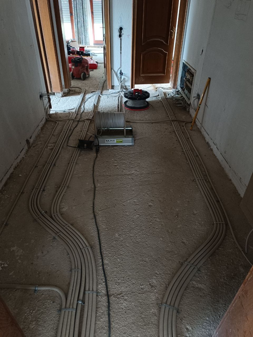 Korytarz w trakcie prac elektrycznych: liczne rury instalacyjne ułożone wzdłuż ścian, sprzęt elektryczny na podłodze, widoczne kable i narzędzia. Surowy stan wykończenia.