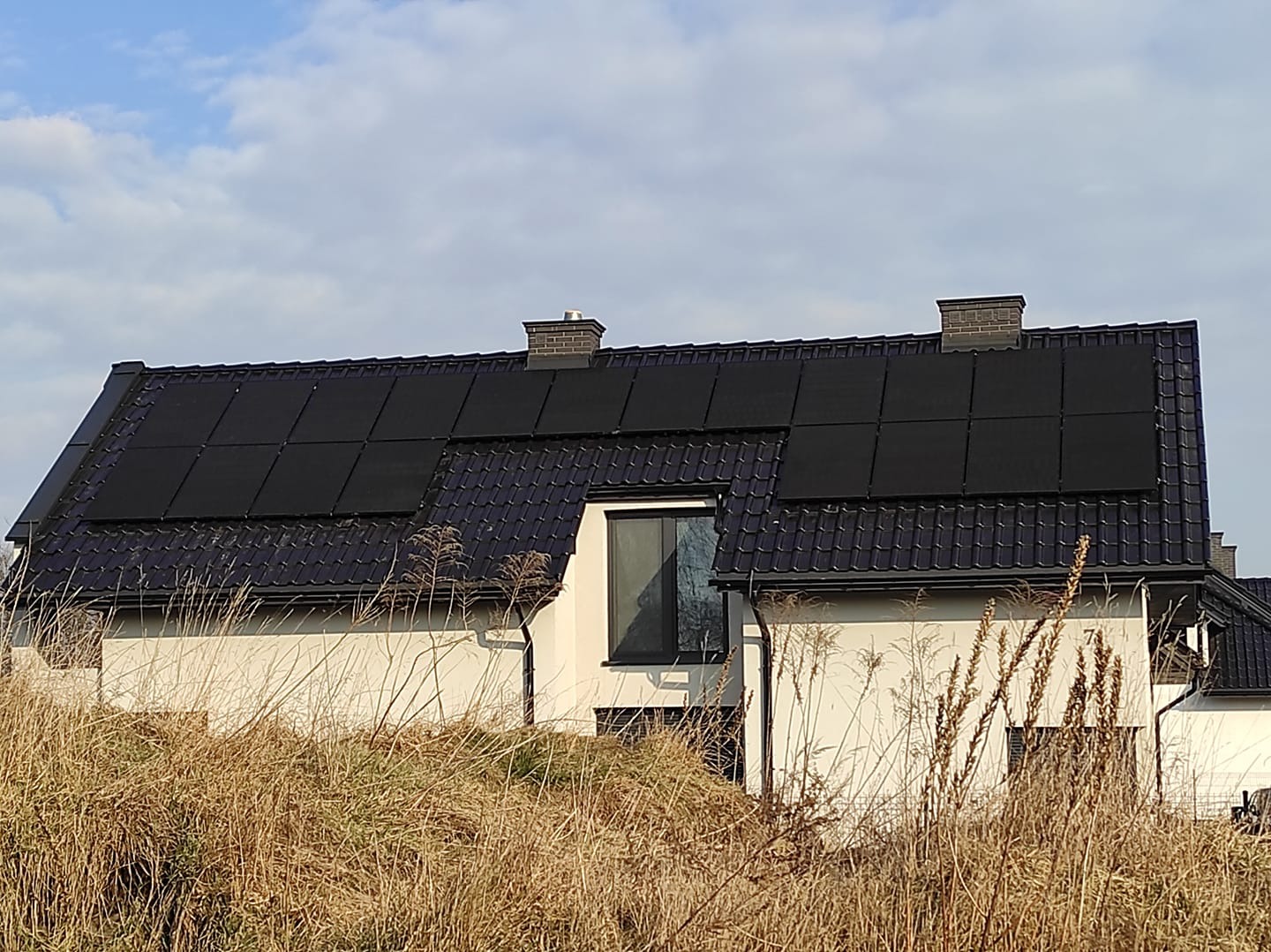 Dom jednorodzinny z czarną dachówką i panelami słonecznymi na dachu, widoczny zza wysokiej trawy. Błękitne niebo w tle.
