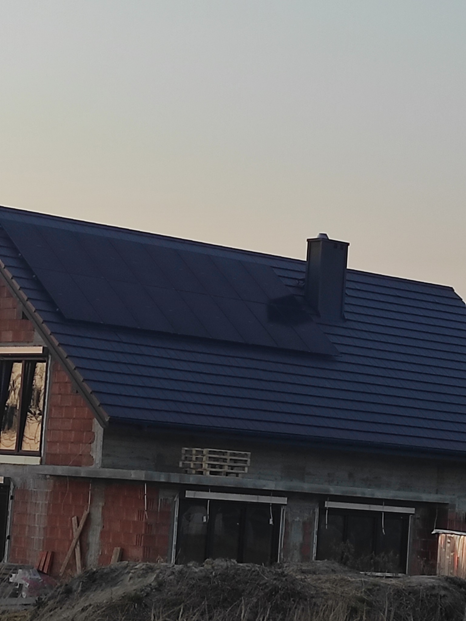 Dom w trakcie budowy z zamontowanymi panelami fotowoltaicznymi na dachu. Widoczne cegły i okna bez wykończenia. Ujęcie z dołu, w tle niebo.