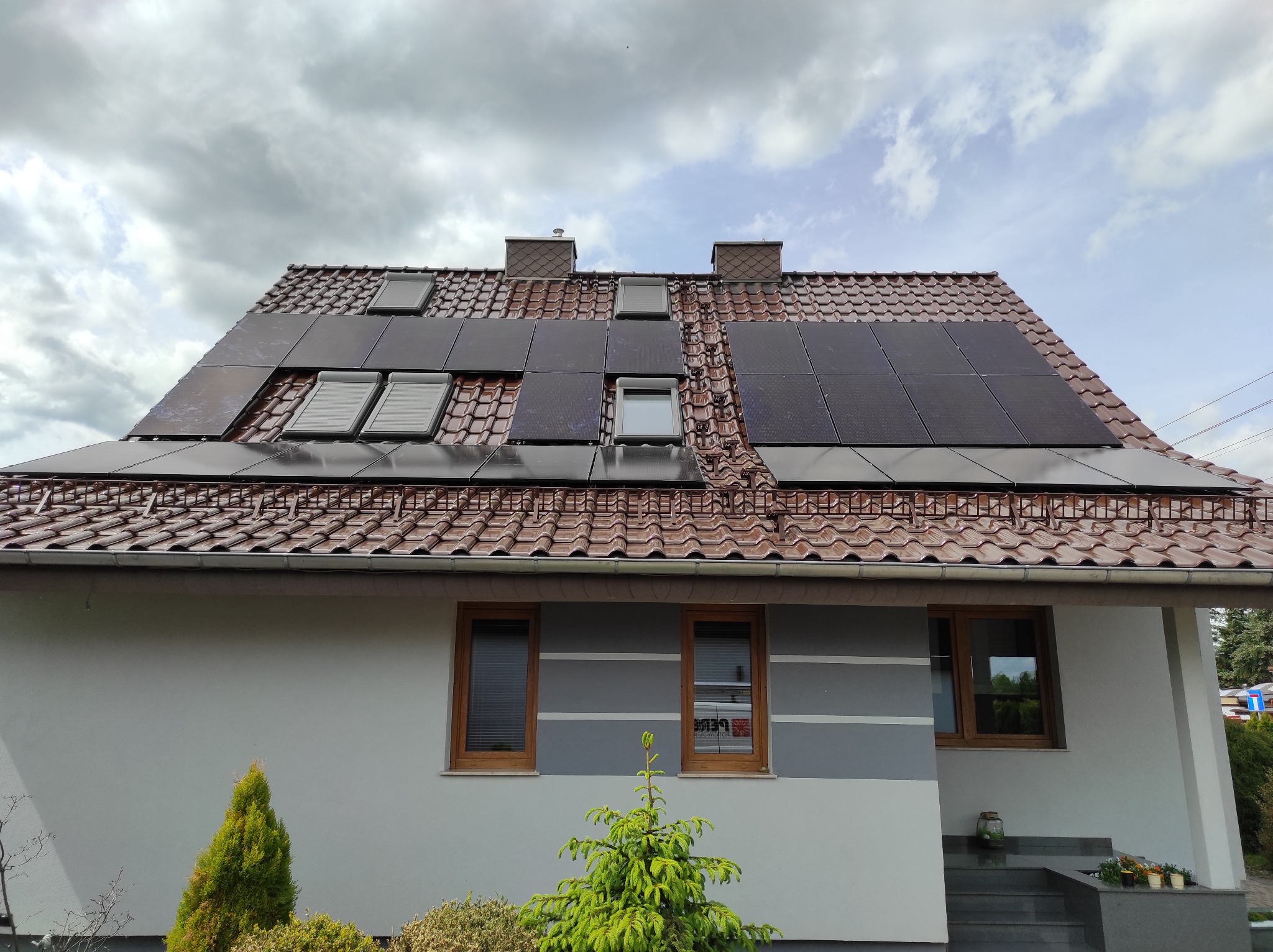 Dom jednorodzinny z panelami fotowoltaicznymi na brązowym dachu. Widoczne okna dachowe i kominy. Szara elewacja z drewnianymi oknami i zielenią.