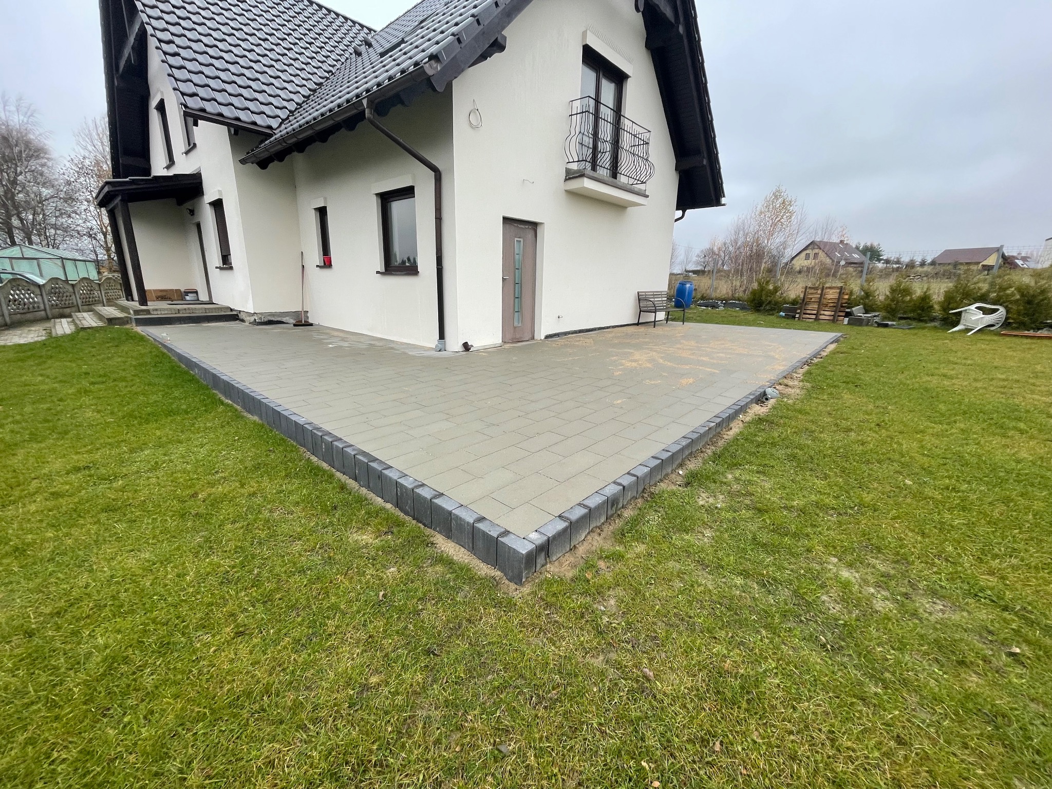 Nowoczesny taras z kostki brukowej w odcieniach szarości, obrzeża z ciemnych kostek, przylegający do białego domu z czarnym dachem. Widok na zadbany trawnik i zabudowę w tle.