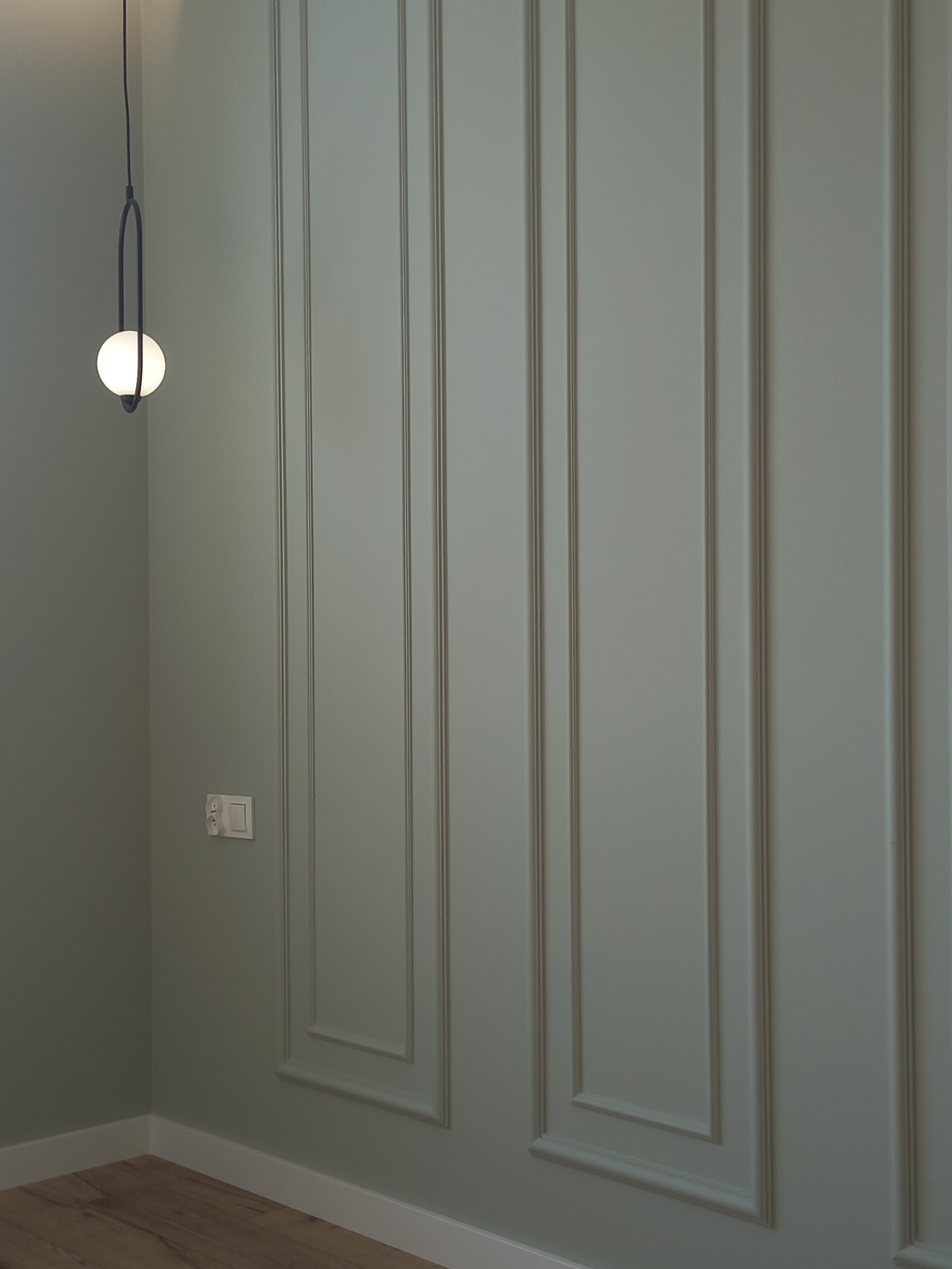 Ściana z dekoracyjnymi panelami w odcieniach szarości, oświetlona nowoczesną lampą wiszącą z okrągłym kloszem. Widoczny fragment podłogi i gniazdko elektryczne.
