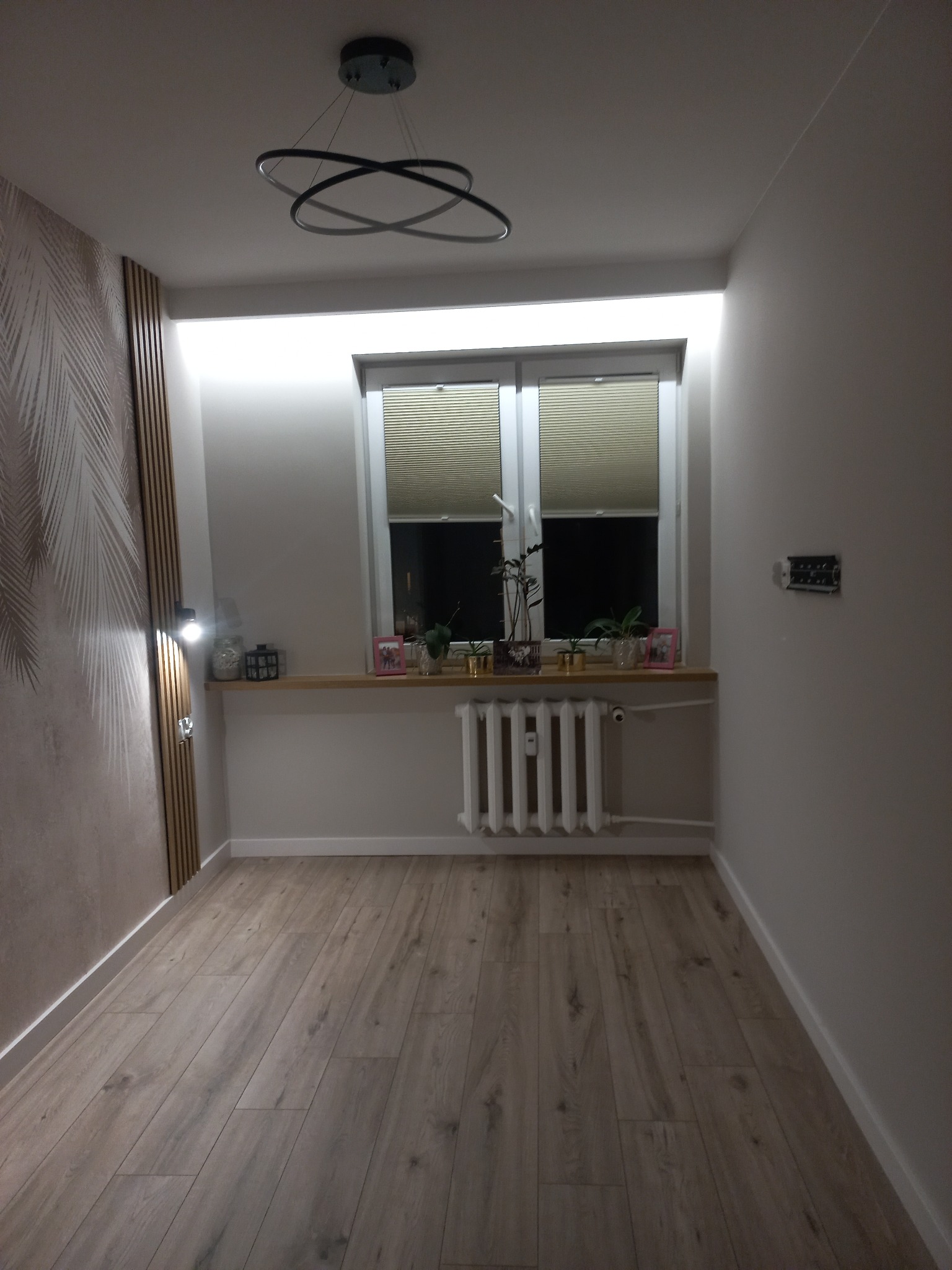 Jasne wnętrze pokoju po remoncie: drewniana podłoga, dekoracyjna ściana z motywem liści, okno z żaluzjami i nowoczesna lampa sufitowa. Widok na grzejnik i parapet z roślinami.