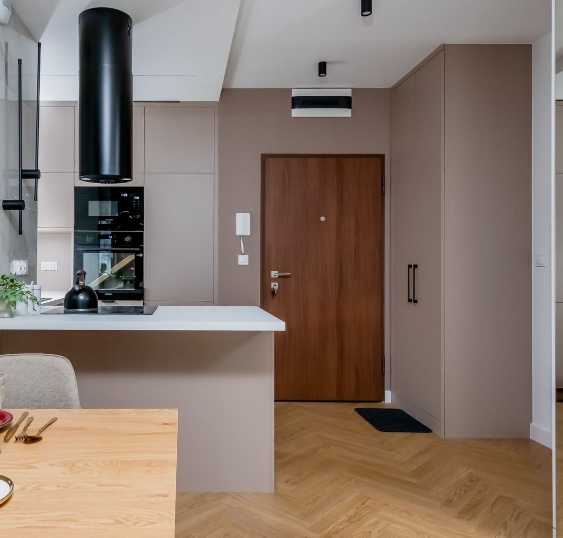 Nowoczesne wnętrze: beżowa zabudowa kuchenna z czarnym okapem cylindrycznym, drewniane drzwi wejściowe i parkiet w jodełkę. Minimalistyczny design, stonowana kolorystyka.