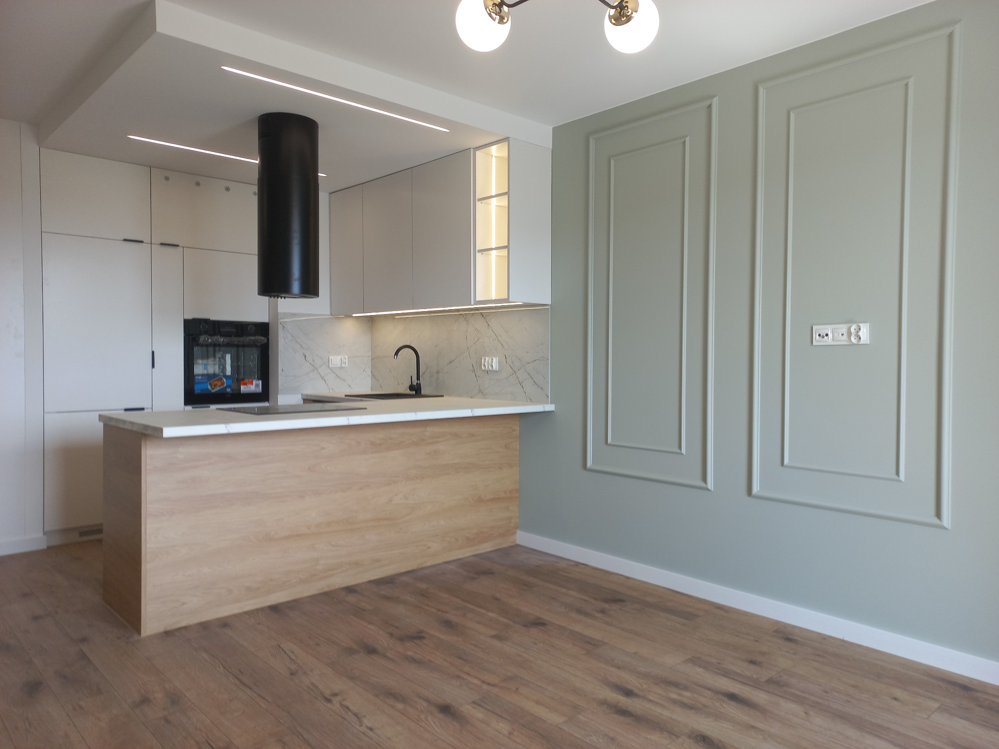 Nowoczesna kuchnia z wyspą i marmurowym blatem, minimalistyczne szafki w odcieniach bieli, czarny okap cylindryczny, ściana z dekoracyjnymi panelami w kolorze pistacjowym.