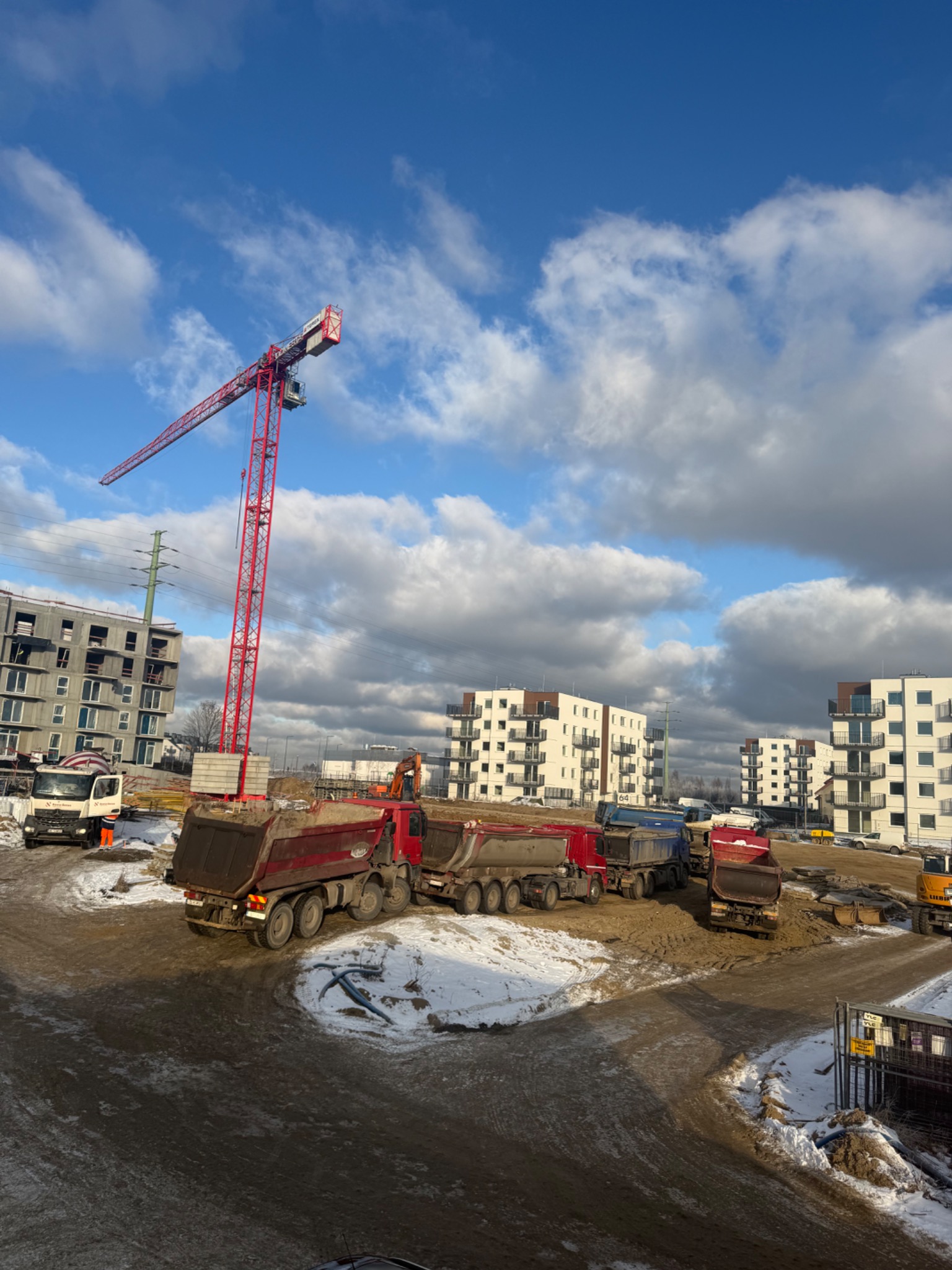 Ciężarówki wywożą ziemię z placu budowy z czerwonym dźwigiem na tle błękitnego nieba z chmurami. W tle bloki mieszkalne w stanie surowym.