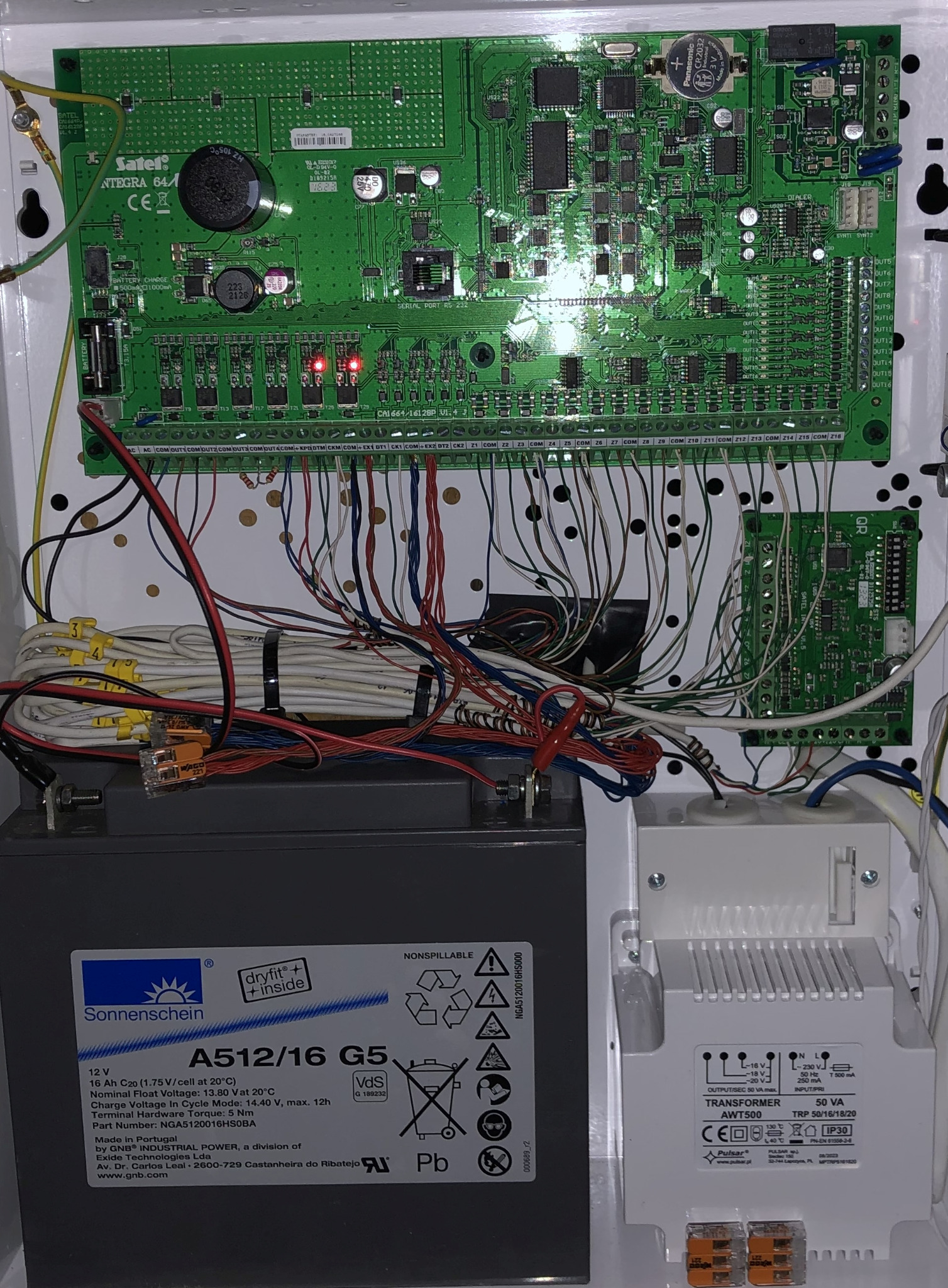 Otwarta obudowa centrali alarmowej Satel Integra 64 z widoczną elektroniką, akumulatorem Sonnenschein i transformatorem Pulsar AWT500. Liczne przewody i złączki.