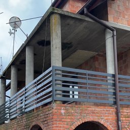 Arkadiusz Nowakowski Usługi Spawalnicze - Szara, pozioma balustrada na ceglanym balkonie z łukami. Widoczne kolumny i anteny na dachu. Budynek w trakcie budowy, surowy stan.
