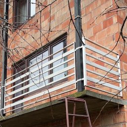 Arkadiusz Nowakowski Usługi Spawalnicze - Balkon z metalową balustradą na tle ceglanej ściany budynku. Do balkonu przystawiona drabina. Widoczne gałęzie drzew. Ujęcie z dołu.