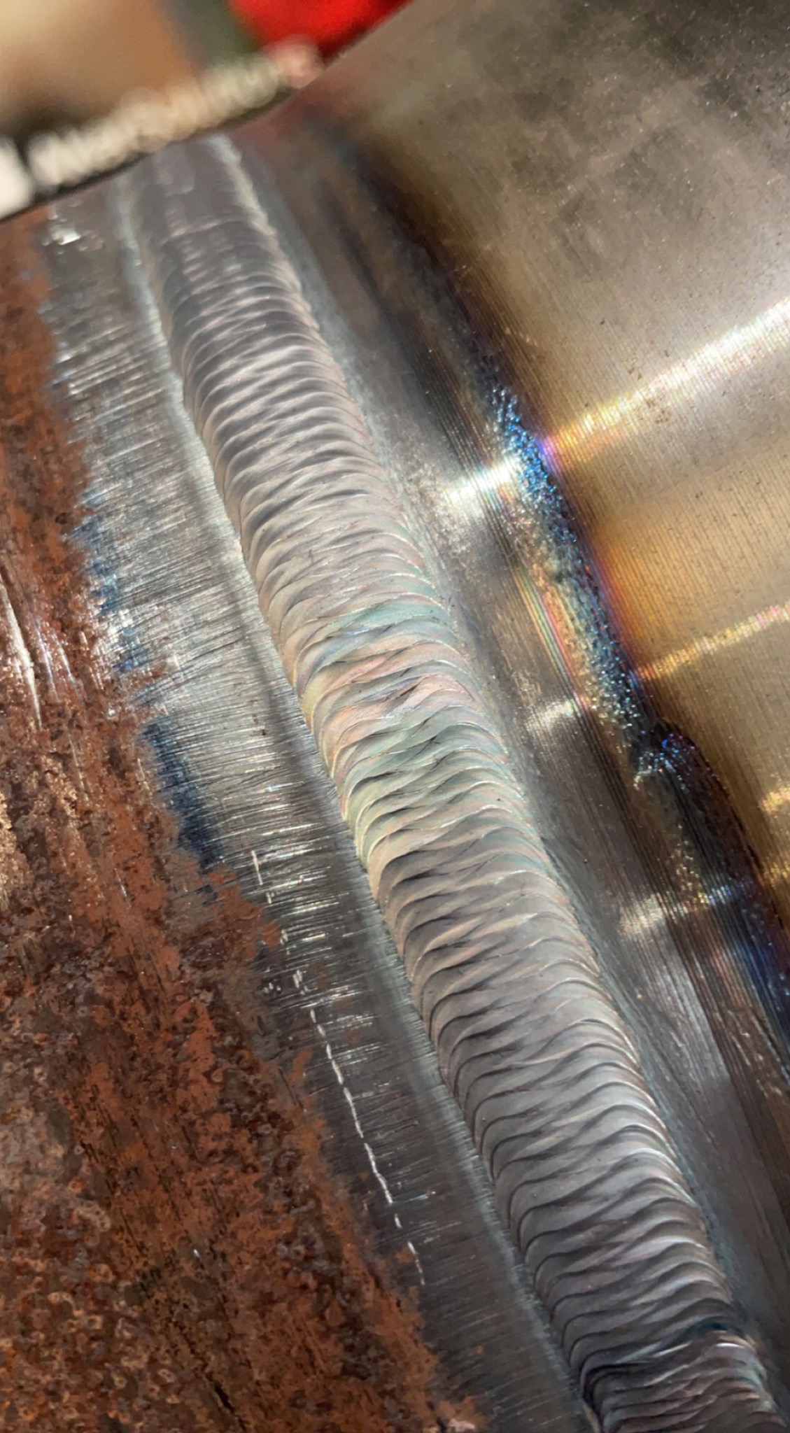 Zbliżenie na świeżo wykonany spaw na metalowej rurze. Widoczna struktura spoiny i przebarwienia termiczne. Część rury pokryta rdzą.