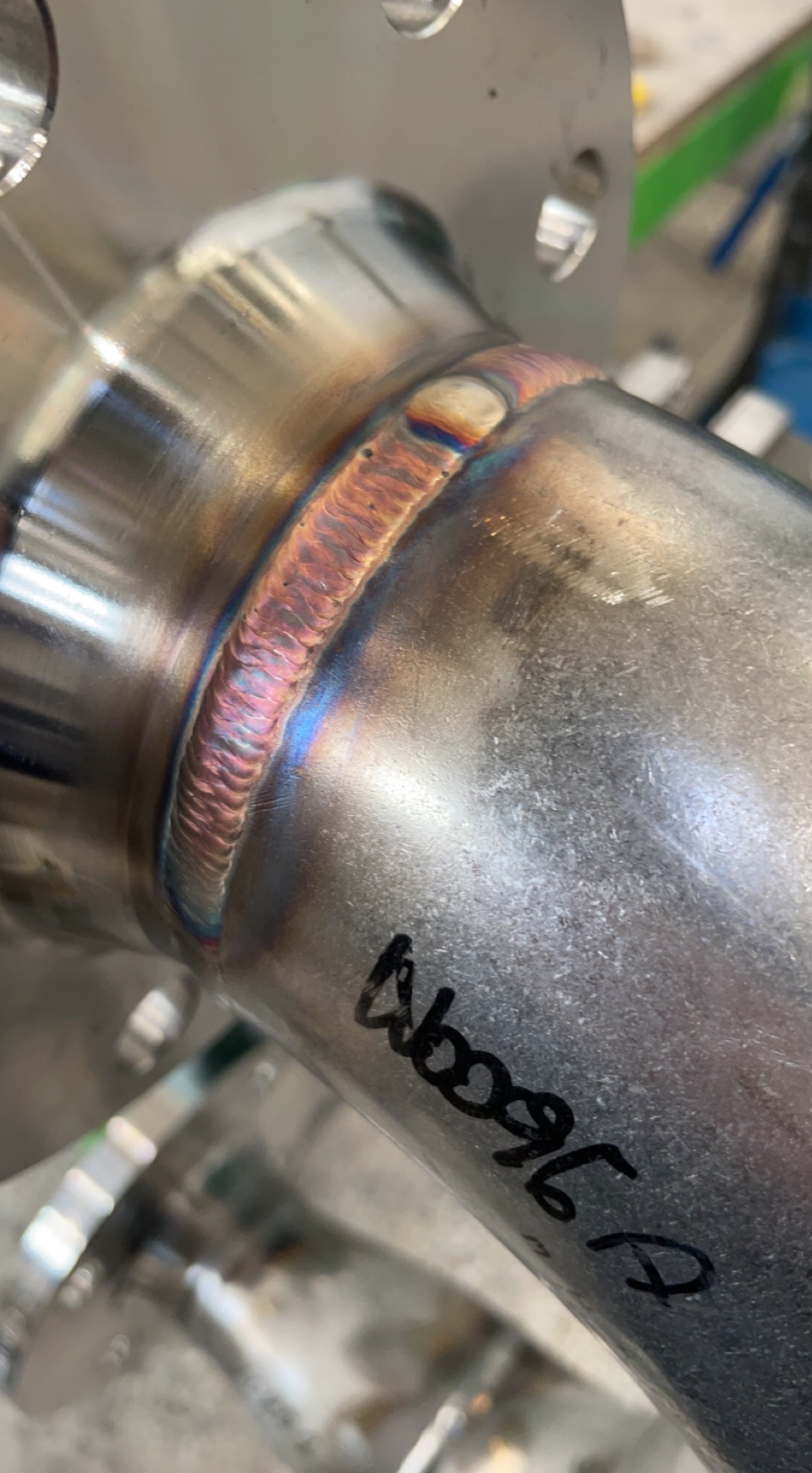 Precyzyjne spawy na metalowej rurze, widoczne tęczowe przebarwienia po spawaniu. Na rurze widnieje odręczny napis. Detal z bliska.
