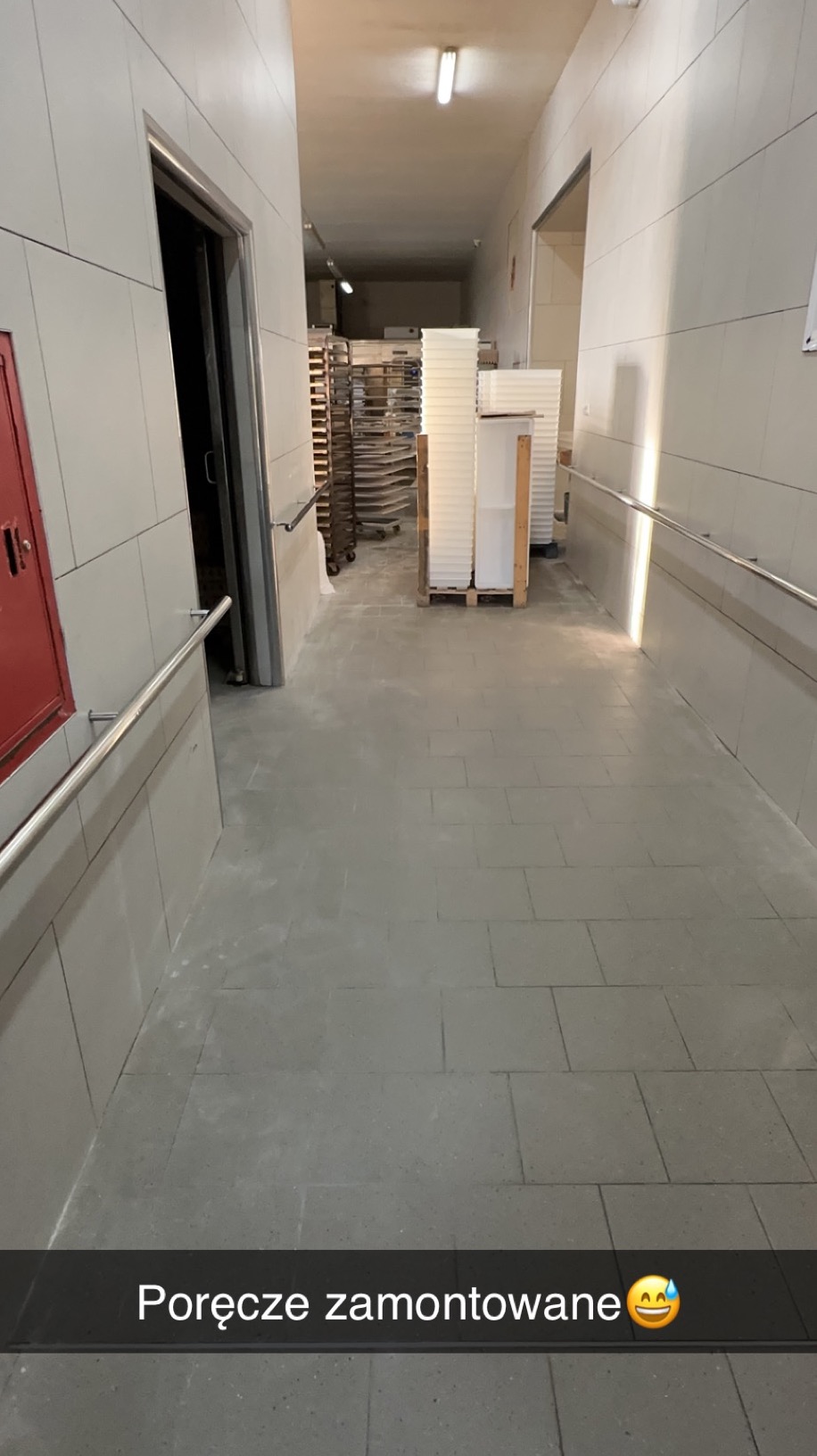 Zamontowane, metalowe poręcze wzdłuż korytarza wyłożonego szarymi płytkami, prowadzącego do pomieszczenia z regałami i pojemnikami. Widok z perspektywy osoby stojącej.