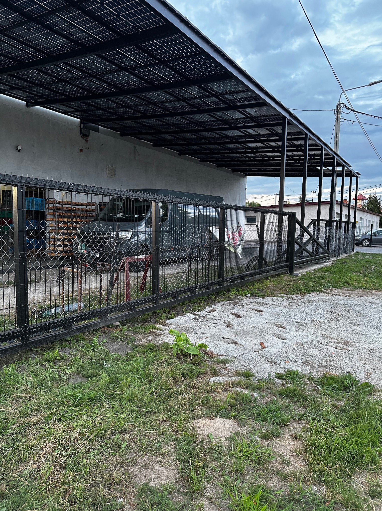 Nowoczesne, czarne ogrodzenie z siatki i metalowa konstrukcja z panelami solarnymi nad wjazdem, widoczne z zewnątrz na tle budynku i zieleni.
