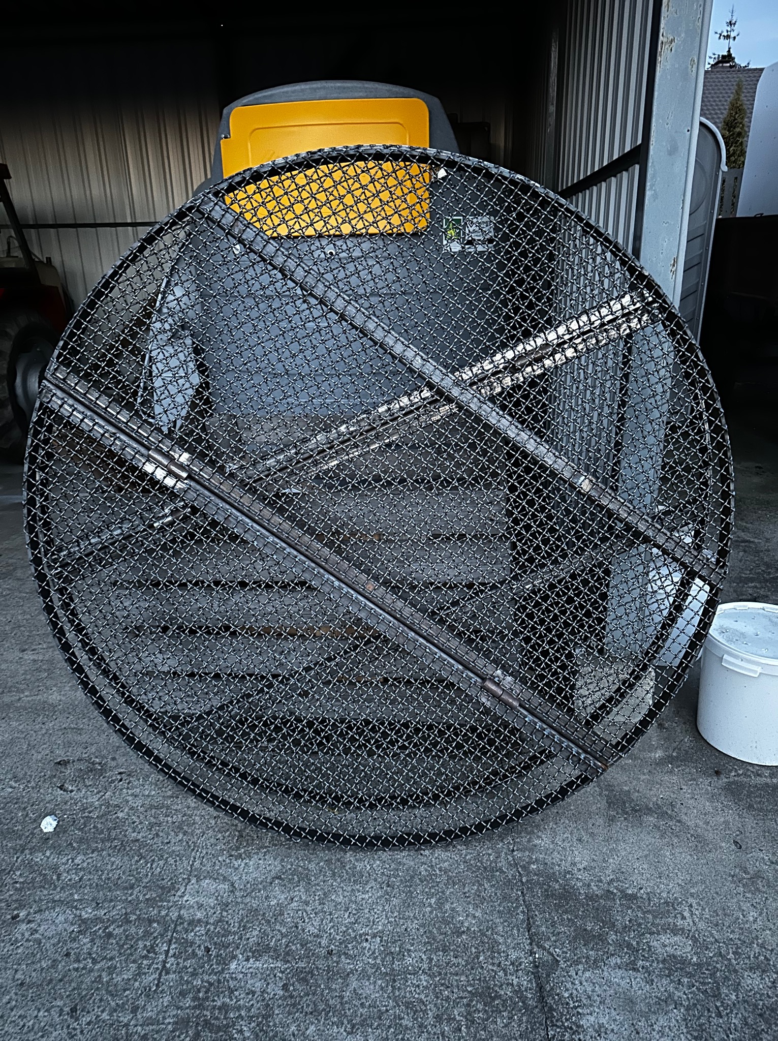 Okrągła konstrukcja z metalowej siatki z poprzecznymi wzmocnieniami, leżąca na betonowej powierzchni, z widocznym w tle sprzętem budowlanym i elementami otoczenia.