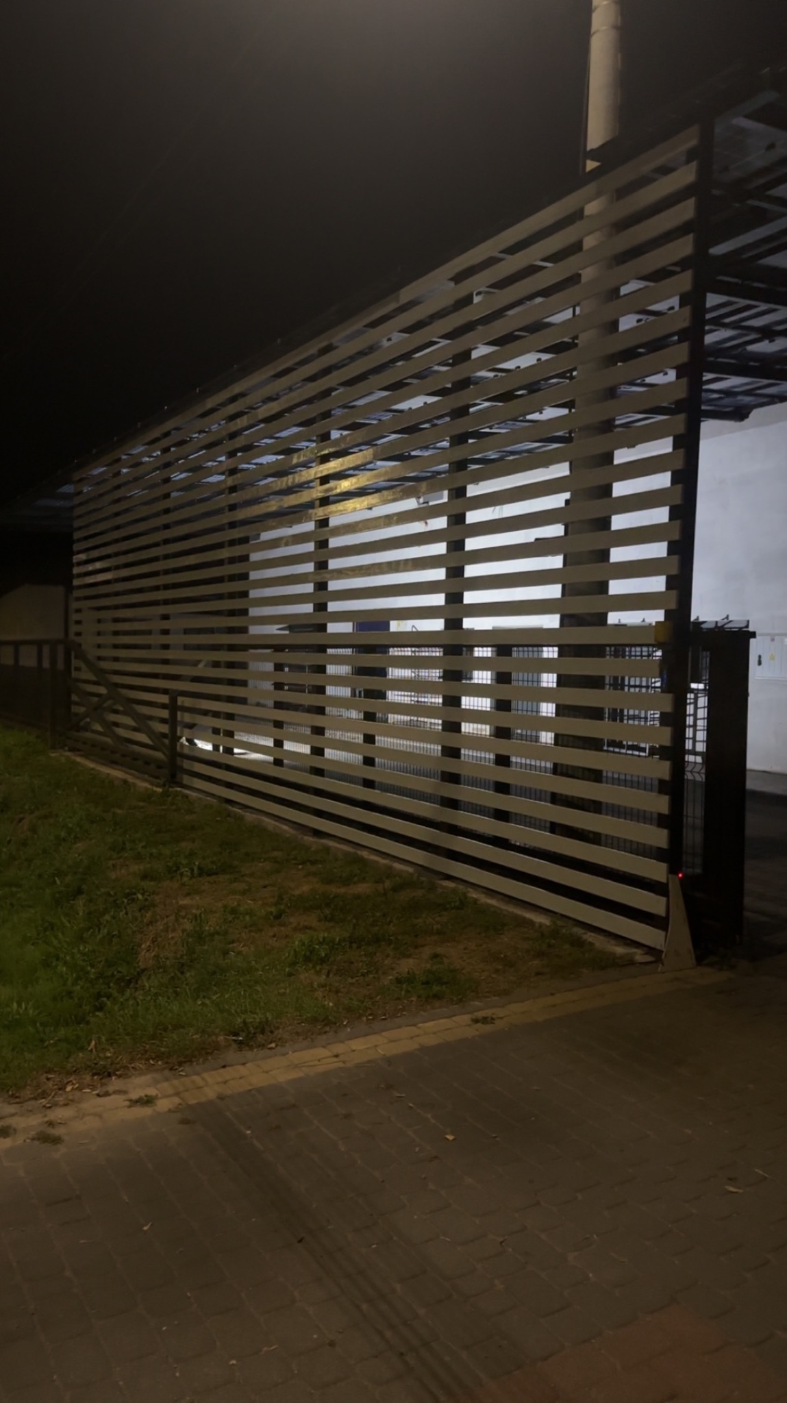 Nowoczesne, poziome przęsła ogrodzeniowe z metalu, widziane nocą. Elementy konstrukcyjne w ciemnym kolorze, zarys zieleni i kostki brukowej w dolnej części kadru.