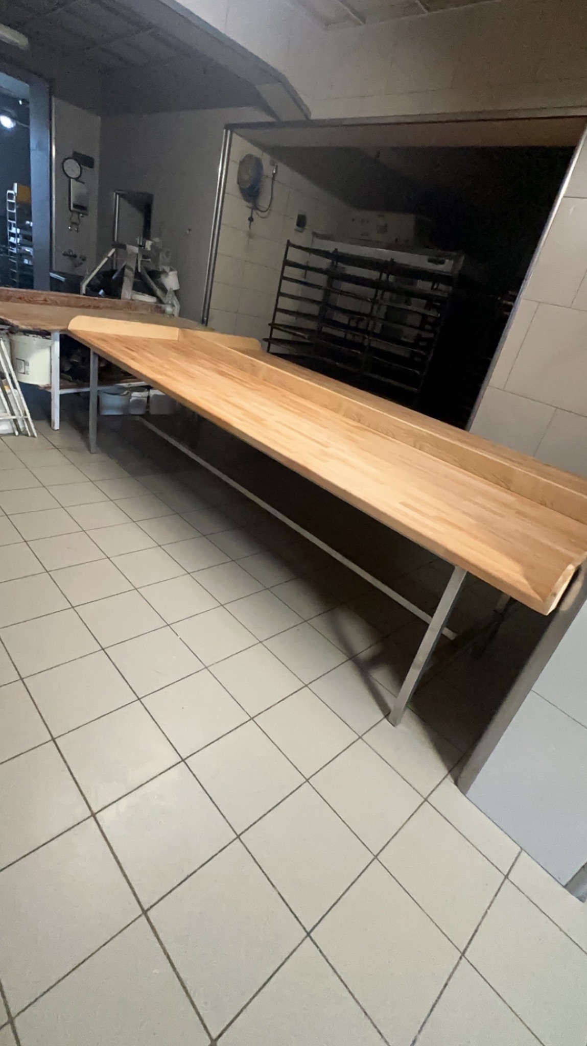 Długi, drewniany stół roboczy na metalowych nogach w pomieszczeniu z wyłożonymi kafelkami ścianami i podłogą. W tle widoczne regały i sprzęt.