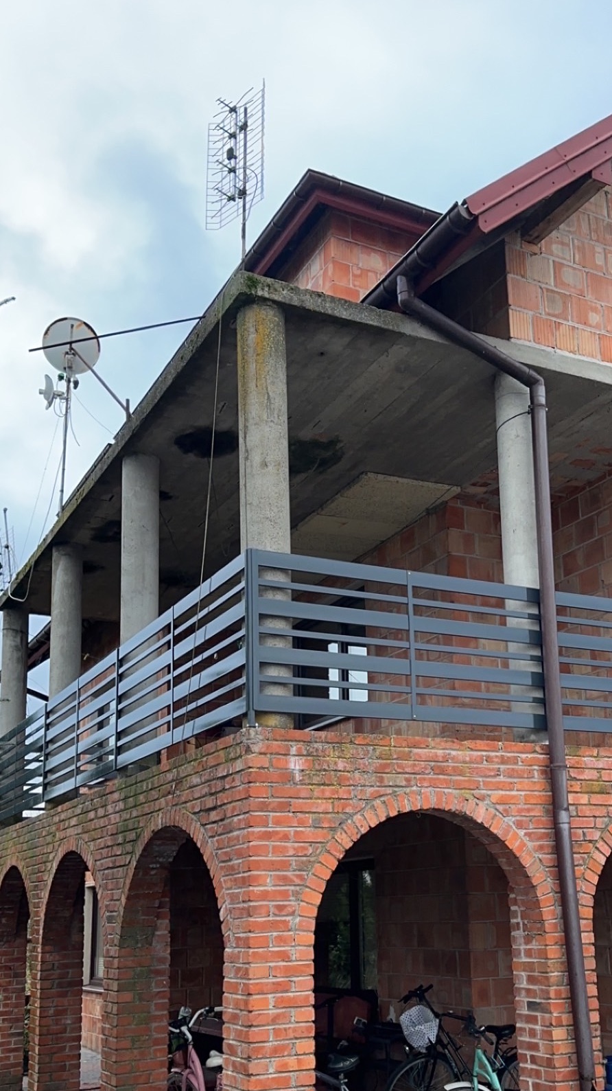 Szara, pozioma balustrada na ceglanym balkonie z łukami. Widoczne kolumny i anteny na dachu. Budynek w trakcie budowy, surowy stan.