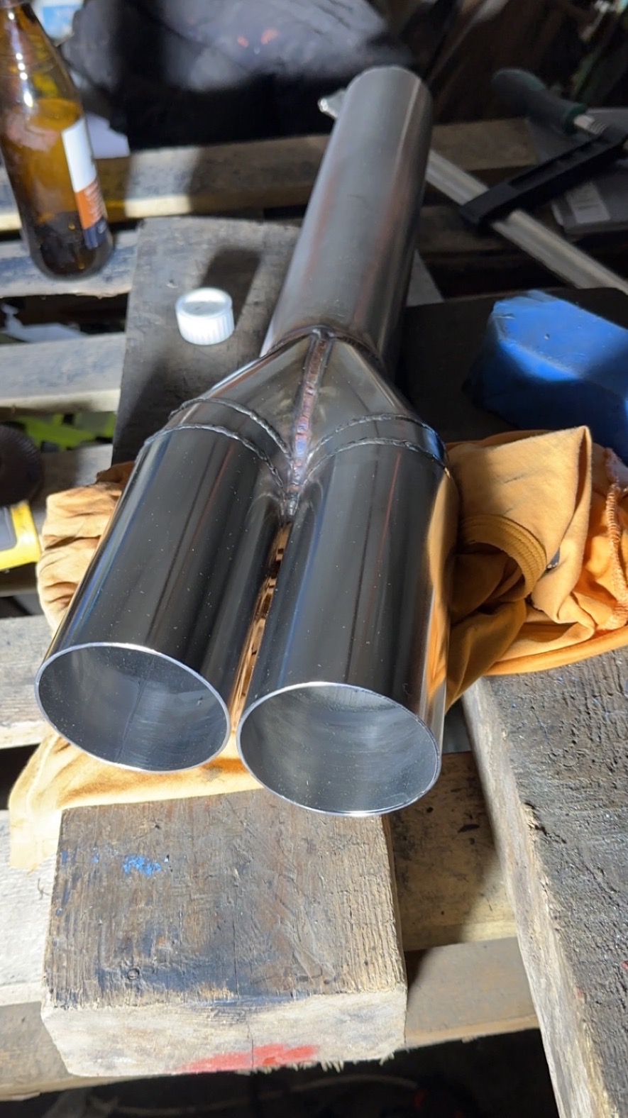 Błyszczący, spawany element metalowy z dwoma wylotami, leżący na drewnianej palecie. Widoczne ślady spawania i otoczenie warsztatowe.