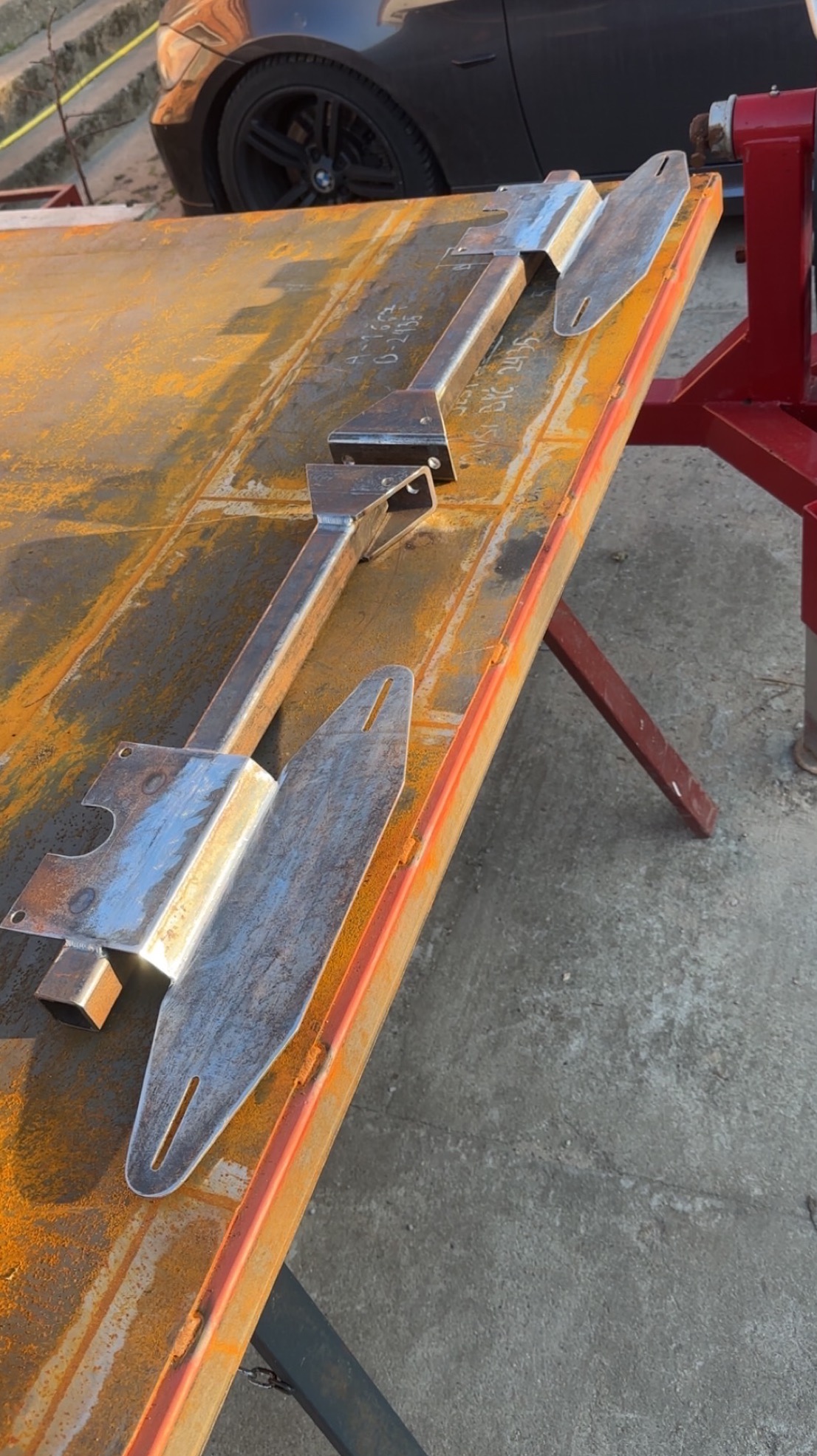 Dwa metalowe elementy konstrukcyjne, prawdopodobnie wsporniki, leżą na zardzewiałym stole warsztatowym. Widoczne ślady spawania i obróbki metalu. W tle fragment samochodu.