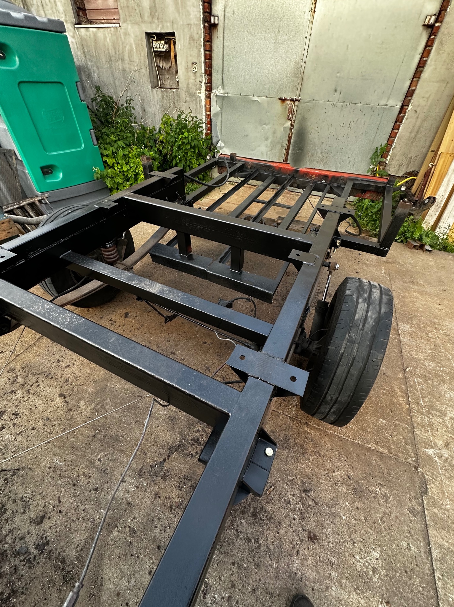 Czarna, metalowa rama przyczepy z kołami, świeżo pomalowana, na betonowym podłożu. W tle ściana budynku z roślinnością i metalowe drzwi.