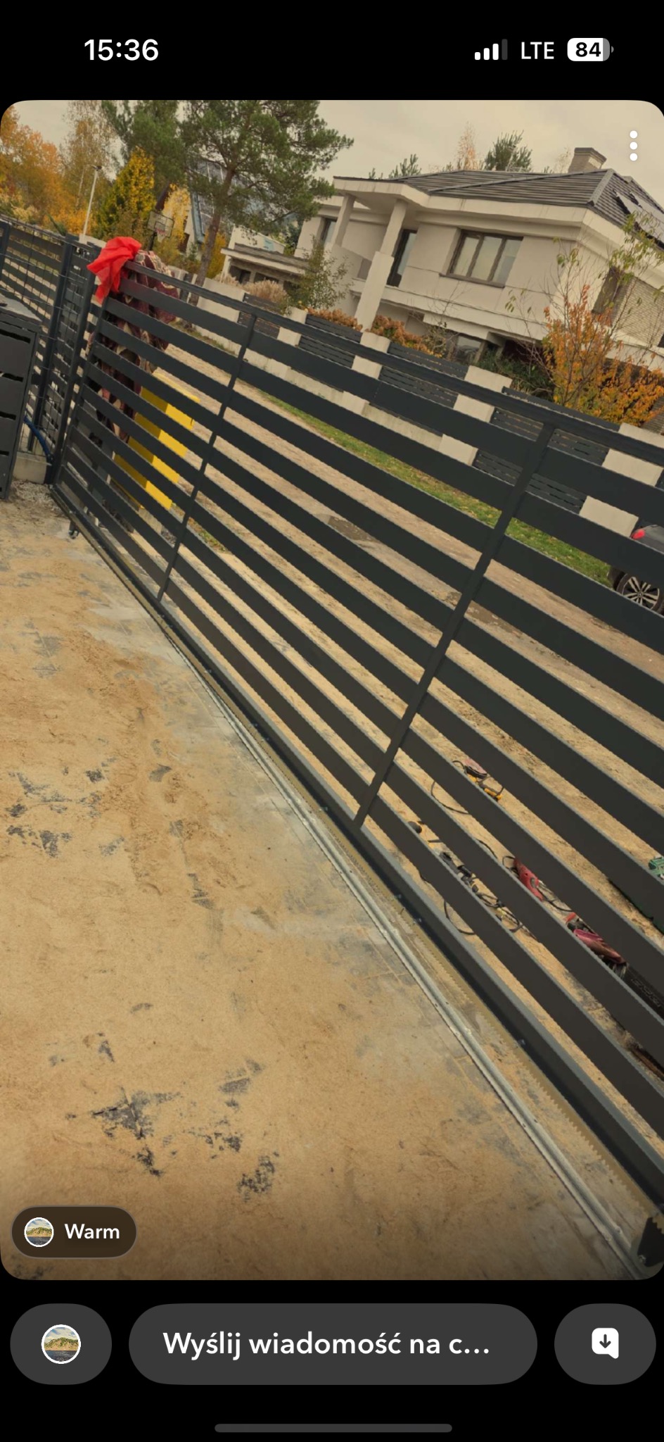 Nowoczesna, pozioma brama wjazdowa z szarych, metalowych profili na tle domu jednorodzinnego i jesiennego krajobrazu. Widoczne narzędzia i elementy montażowe.