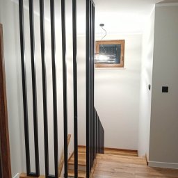 Metalek Kamil Talkowski - Nowoczesna balustrada schodowa z czarnych, pionowych profili, drewniane schody, jasne ściany, okno z drewnianą ramą i lampa wisząca. Minimalistyczny design.