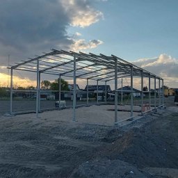 Metalek Kamil Talkowski - Konstrukcja stalowa hali w trakcie montażu, widok z zewnątrz na tle zachodzącego słońca i pochmurnego nieba. Widoczne elementy mocujące i teren budowy.