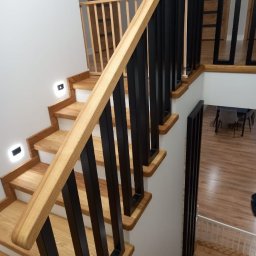 Metalek Kamil Talkowski - Nowoczesne schody z drewnianymi stopniami i czarną, metalową balustradą. Widoczne oświetlenie schodowe i fragment wnętrza domu z drewnianą podłogą.