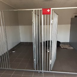 Metalek Kamil Talkowski - Metalowe boksy z kratami w pomieszczeniu, z flagą Szwajcarii na jednym z nich. Widoczne miski i ciemny obiekt wewnątrz jednego z boksów. Szara podłoga i ściany.