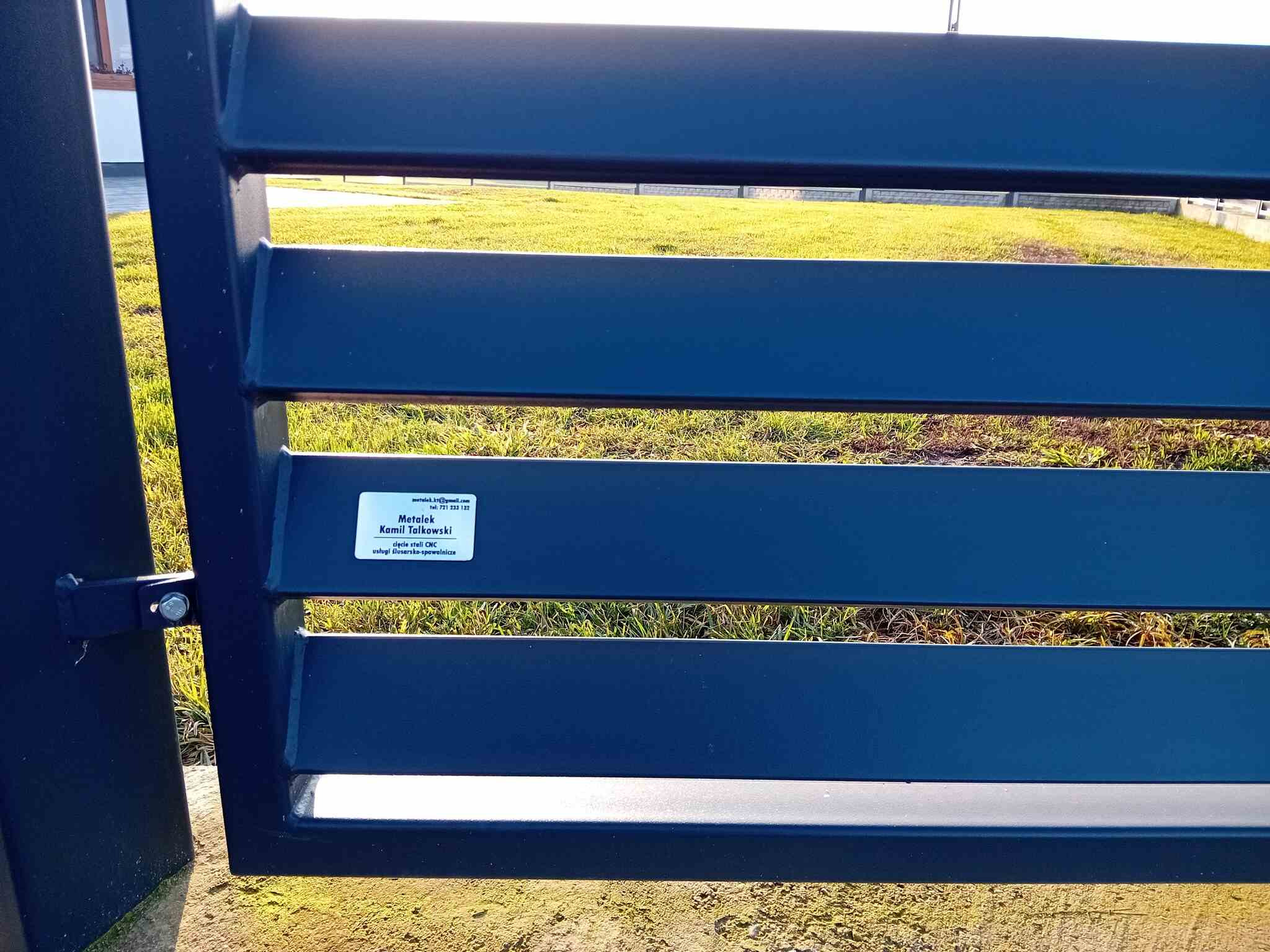 Nowoczesna, pozioma brama wjazdowa z metalu w kolorze ciemnoniebieskim, z naklejką firmy Metalek Kamil Talkowski oferującej usługi ślusarsko-spawalnicze i cięcie CNC.