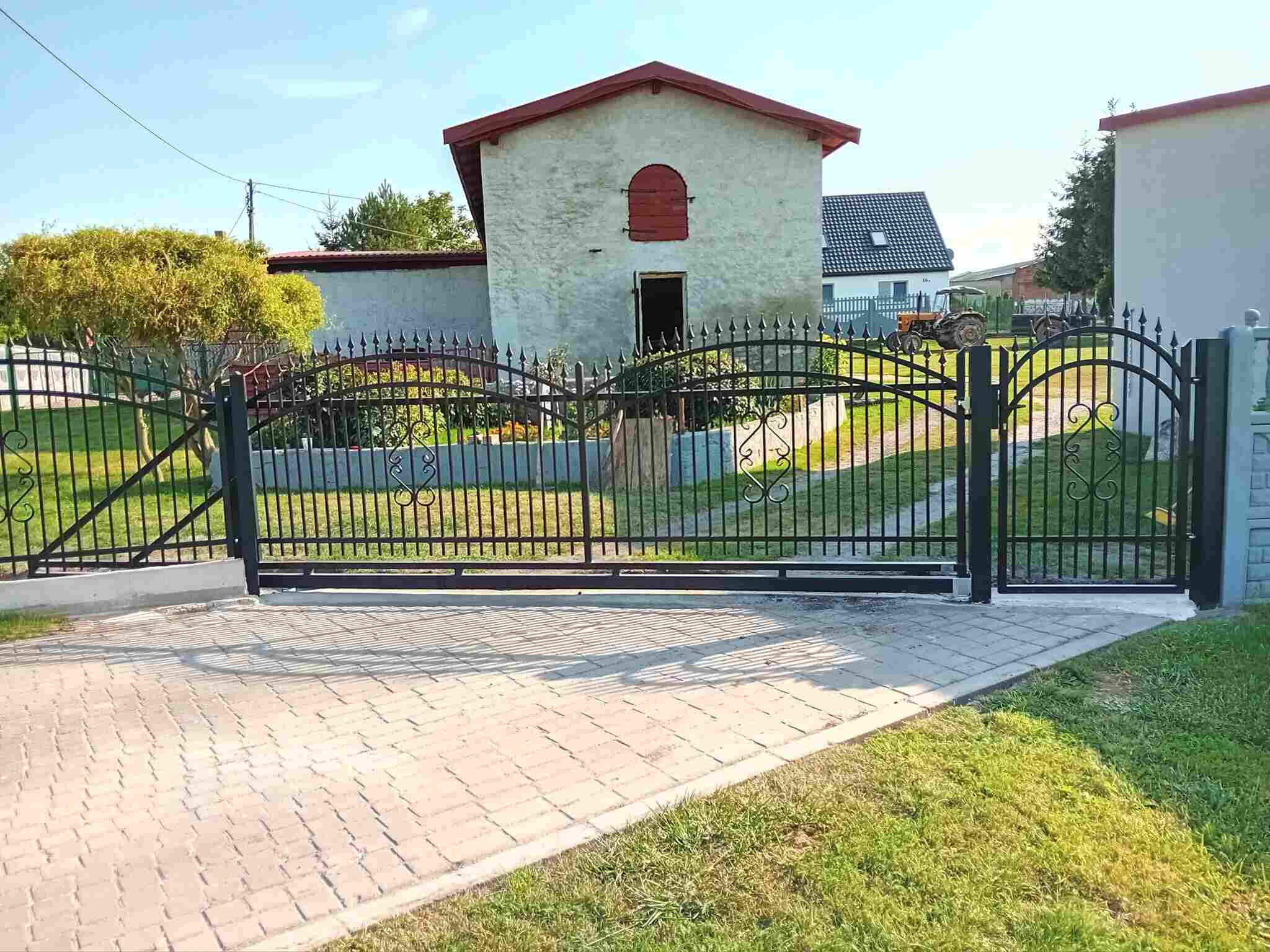 Czarna, metalowa brama wjazdowa z dekoracyjnymi elementami na posesji z budynkiem gospodarczym i traktorem w tle, widziana z poziomu kostki brukowej.