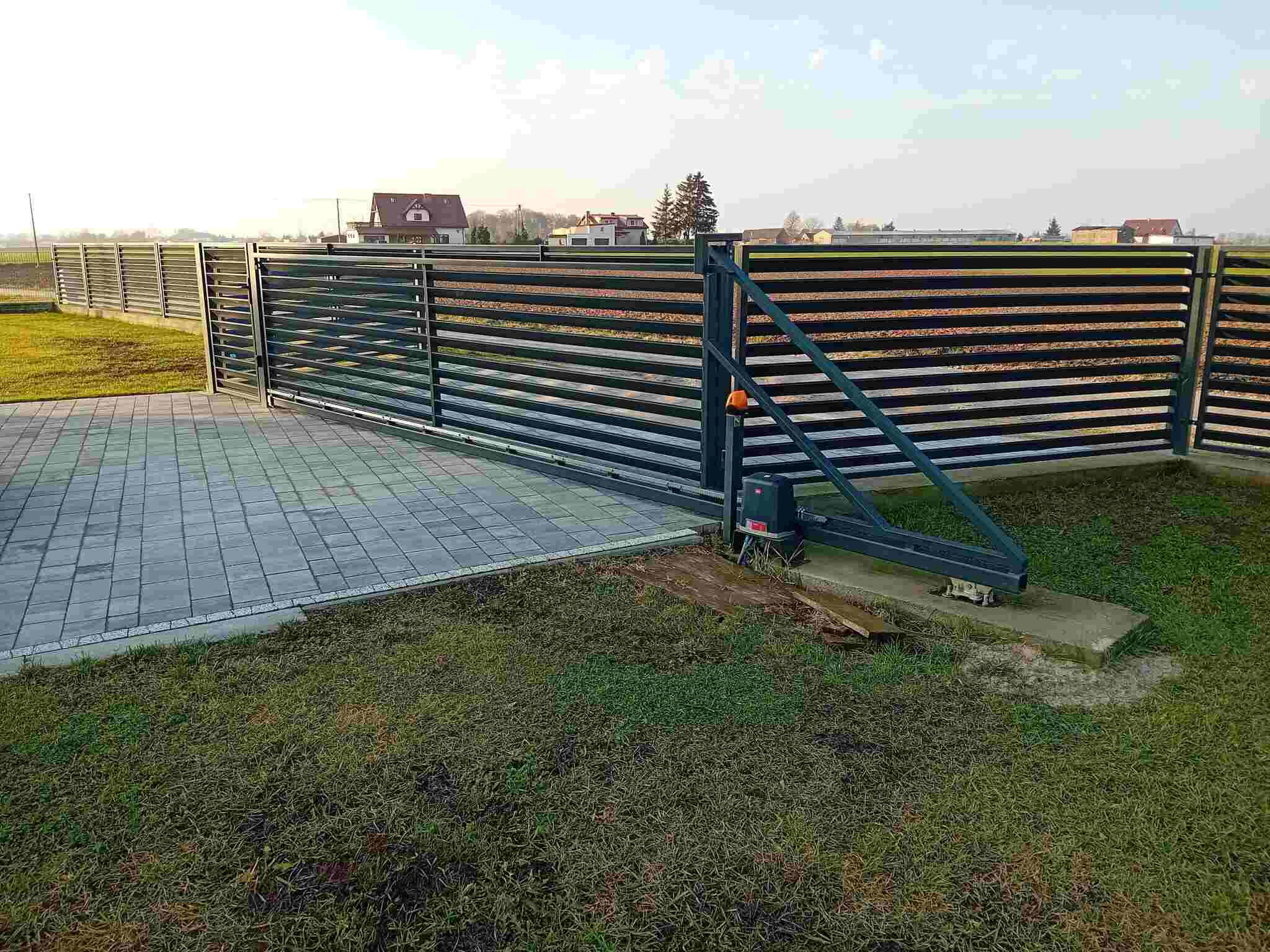 Nowoczesna, przesuwna brama wjazdowa z poziomymi panelami, zautomatyzowana, na tle zielonego trawnika i zabudowań mieszkalnych w oddali. Widoczny mechanizm napędowy.