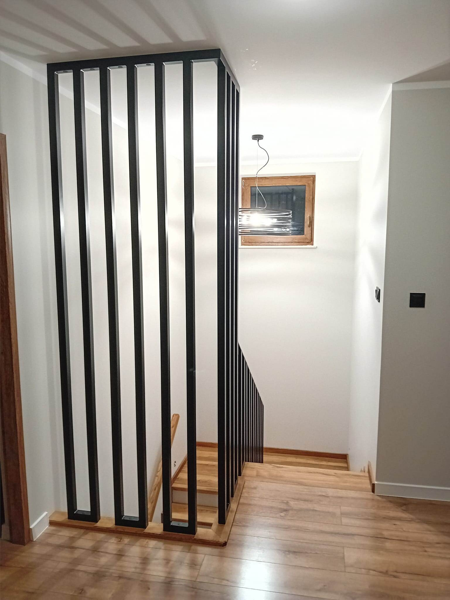 Nowoczesna balustrada schodowa z czarnych, pionowych profili, drewniane schody, jasne ściany, okno z drewnianą ramą i lampa wisząca. Minimalistyczny design.