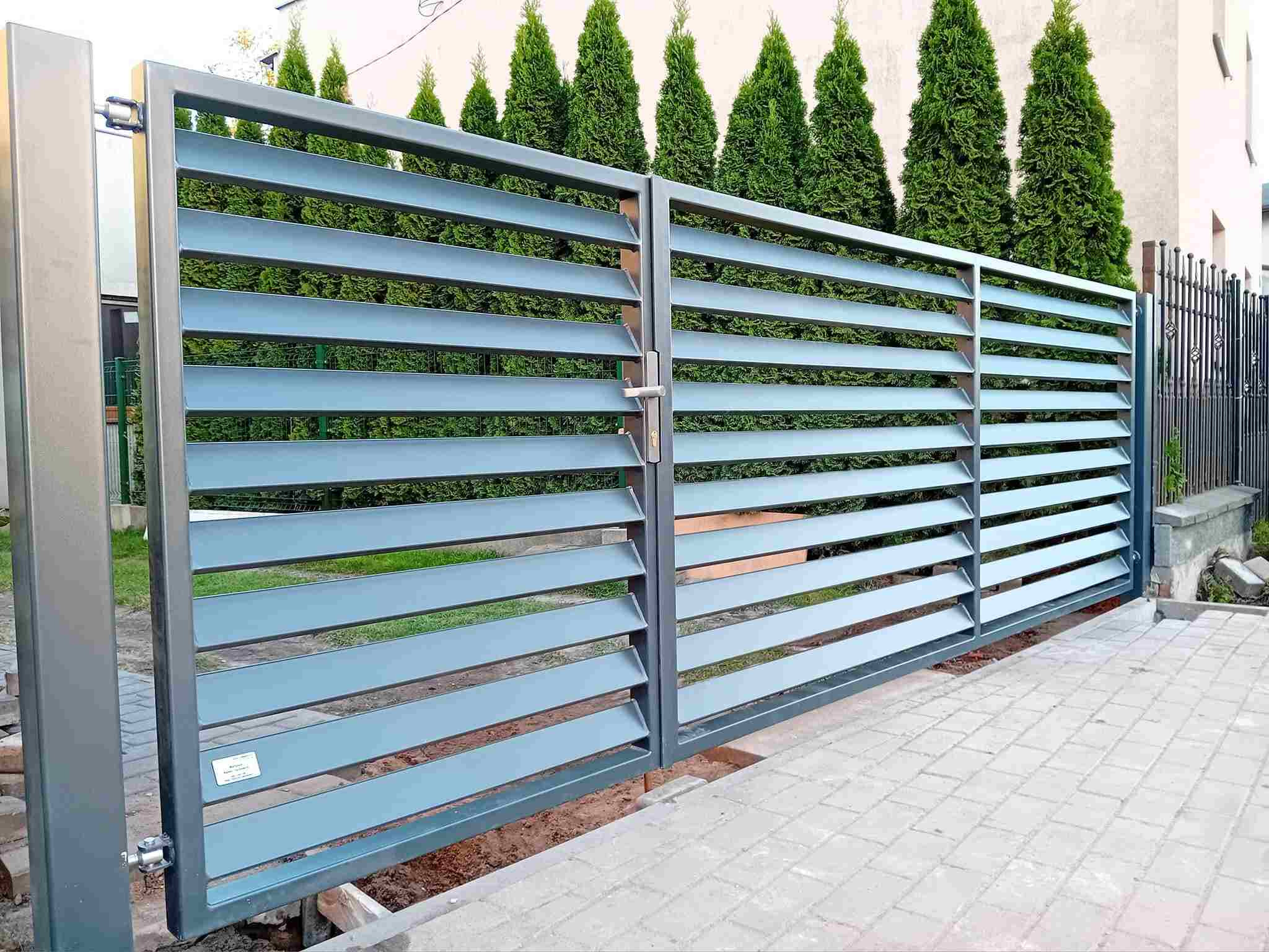Nowoczesna, przesuwna brama wjazdowa z poziomymi lamelami w odcieniach szarości, na tle zielonych tuj i jasnego budynku. Widoczne detale montażowe i kostka brukowa.
